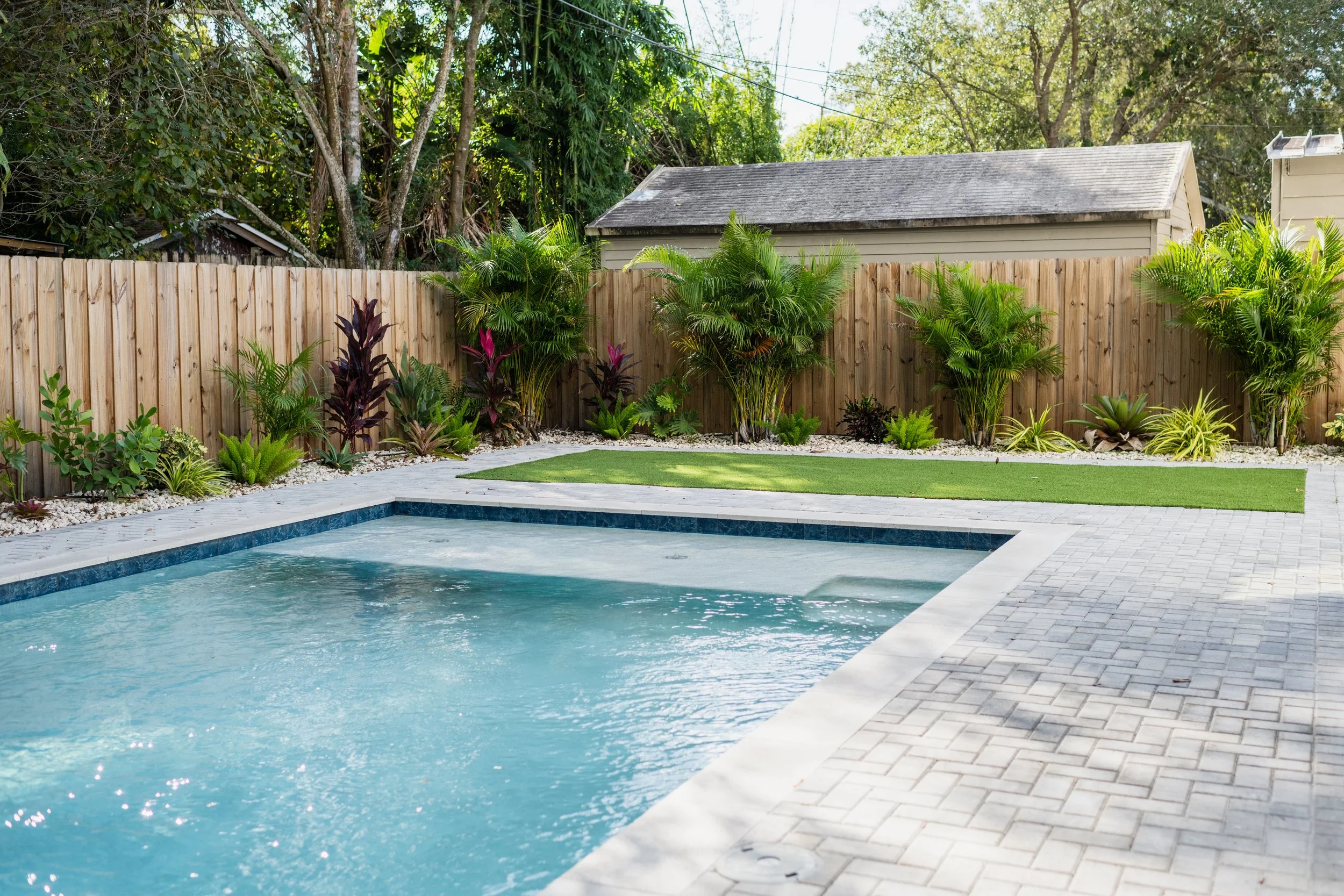 Backyard with an in-ground swimming pool, surrounded by a paved patio, bordered by a wooden fence and lush green plants and shrubs, with trees and a shed in the background.