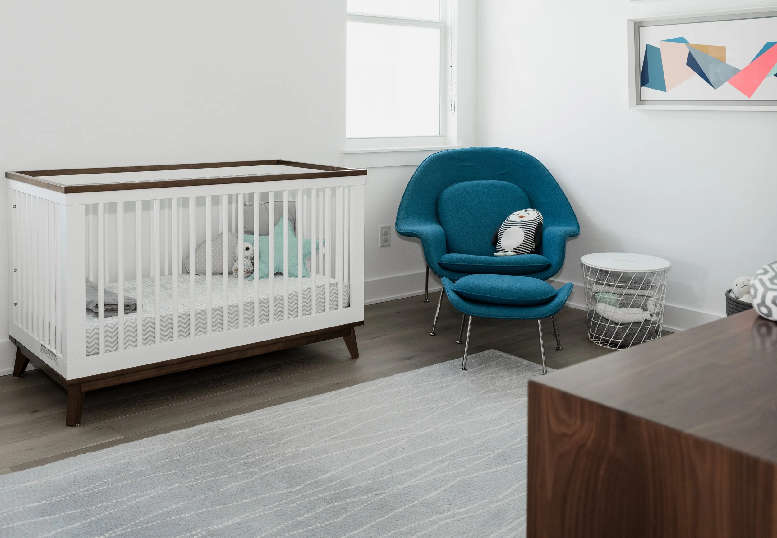 A nursery with a white crib on the left, containing plush toys and stuffed animals. To the right, a blue mid-century modern armchair with a small pillow, next to a white wire basket filled with blankets, and framed abstract art on the white wall.
