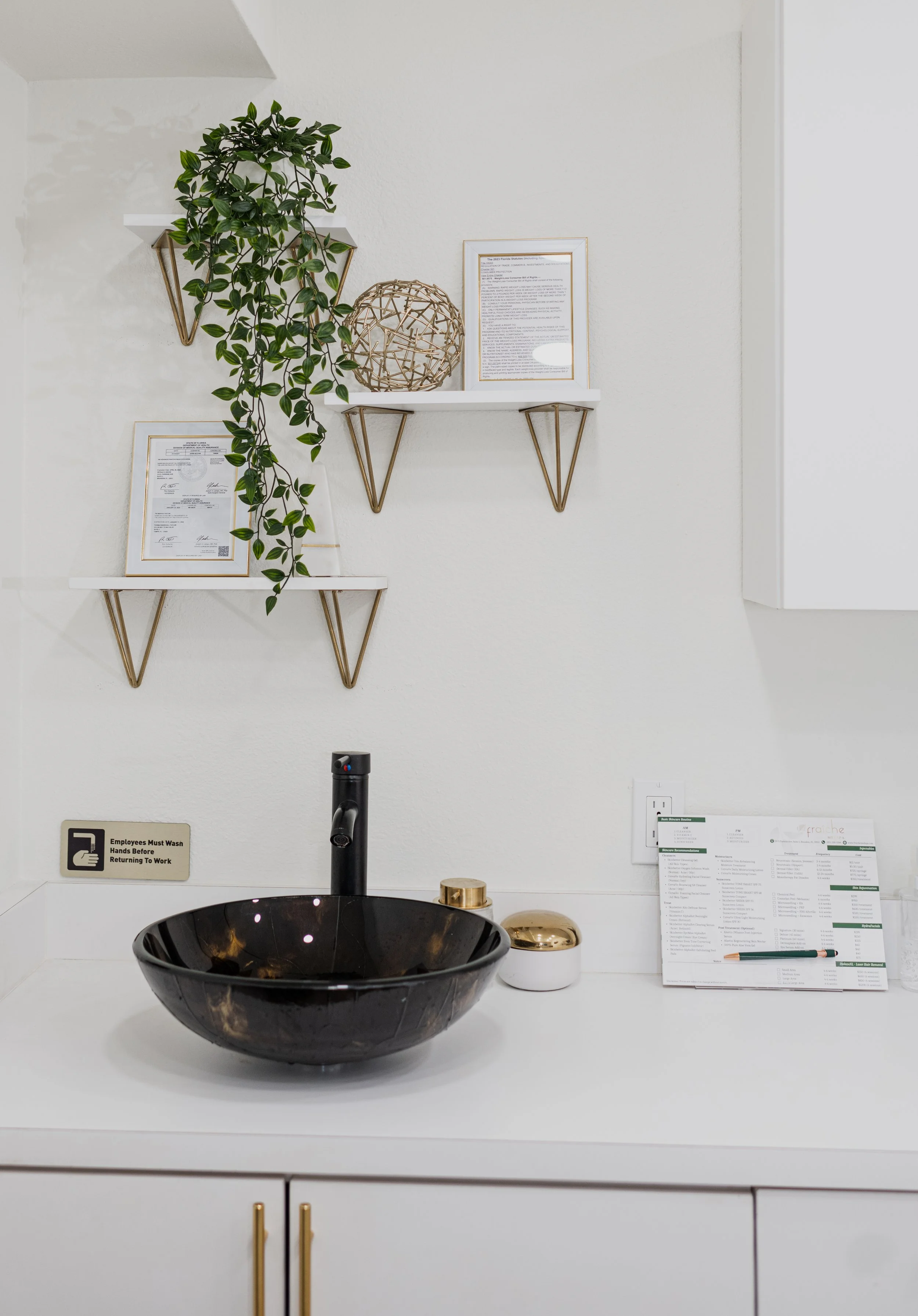 White bathroom vanity with black vessel sink, black faucet, and two small white and gold containers. White wall with three decorative white shelves holding framed papers and a green trailing plant, and a white cabinet. Sign indicating employees must 
