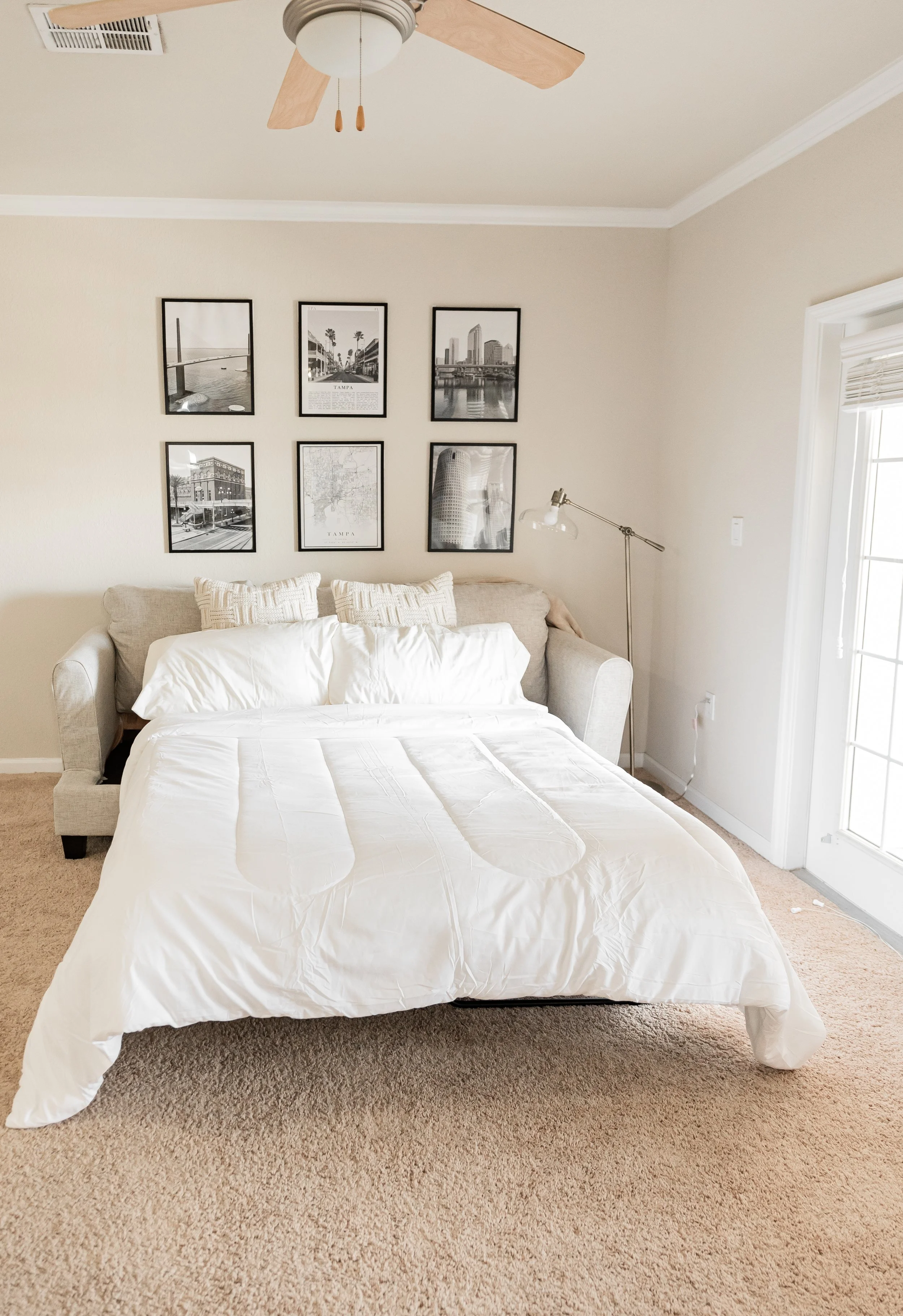A bedroom with a bed covered in white bedding and pillows, a beige sofa at the head of the bed, a wall art gallery above the sofa, a floor lamp next to the sofa, a ceiling fan, a window with blinds, and beige carpet flooring.