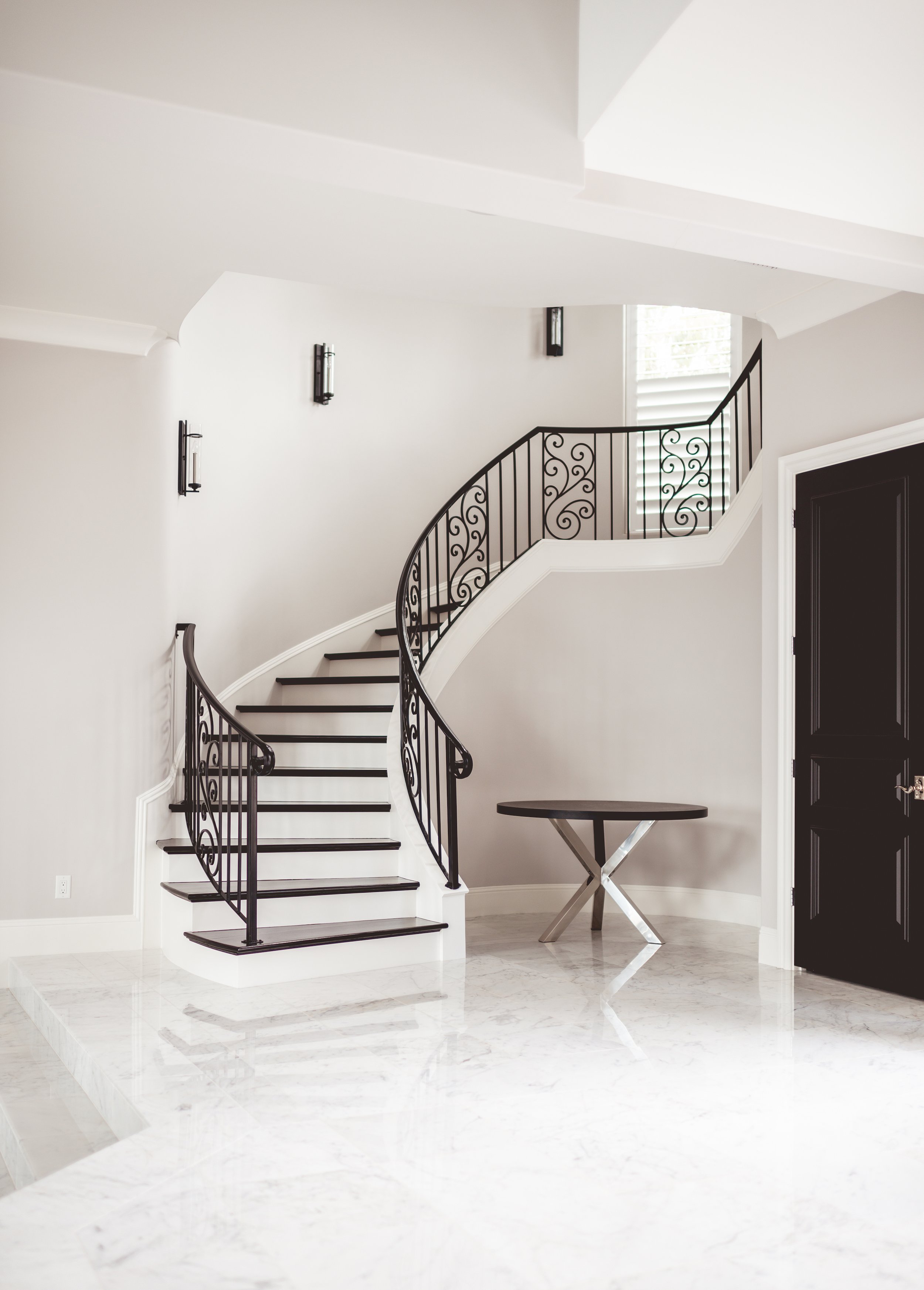 Modern interior with a curved staircase featuring black wrought iron railing, white walls, and marble flooring. There is a small black table beneath a window with white blinds, and three wall-mounted black light fixtures.