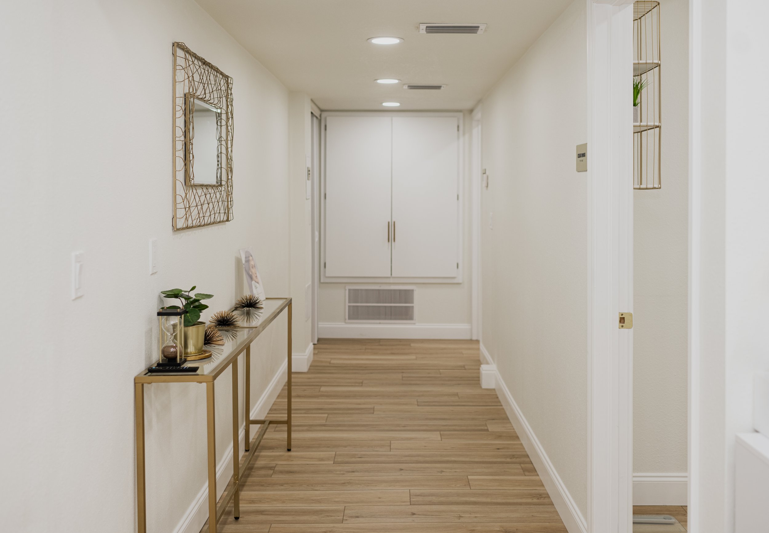A modern hallway with light-colored walls, wooden flooring, and white trim. There is a gold-framed mirror and a gold console table with decorative items on the left side. At the end, double white doors are visible, along with a vent below them. There
