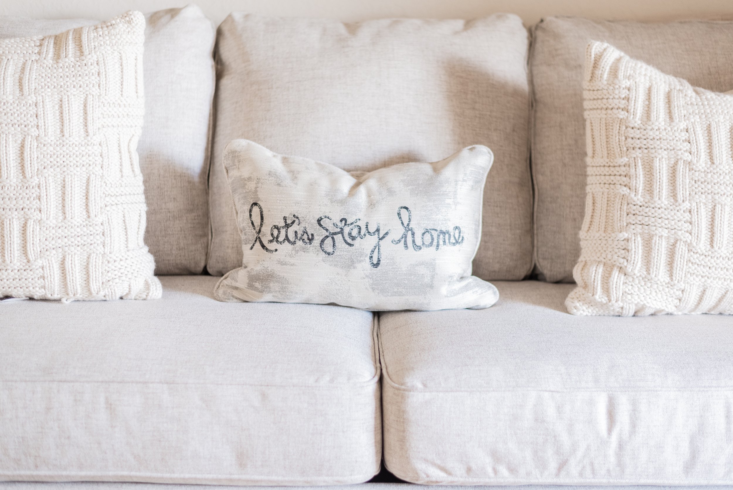 A light-colored sofa with three decorative pillows, including one with the words "let's stay home" written in cursive.