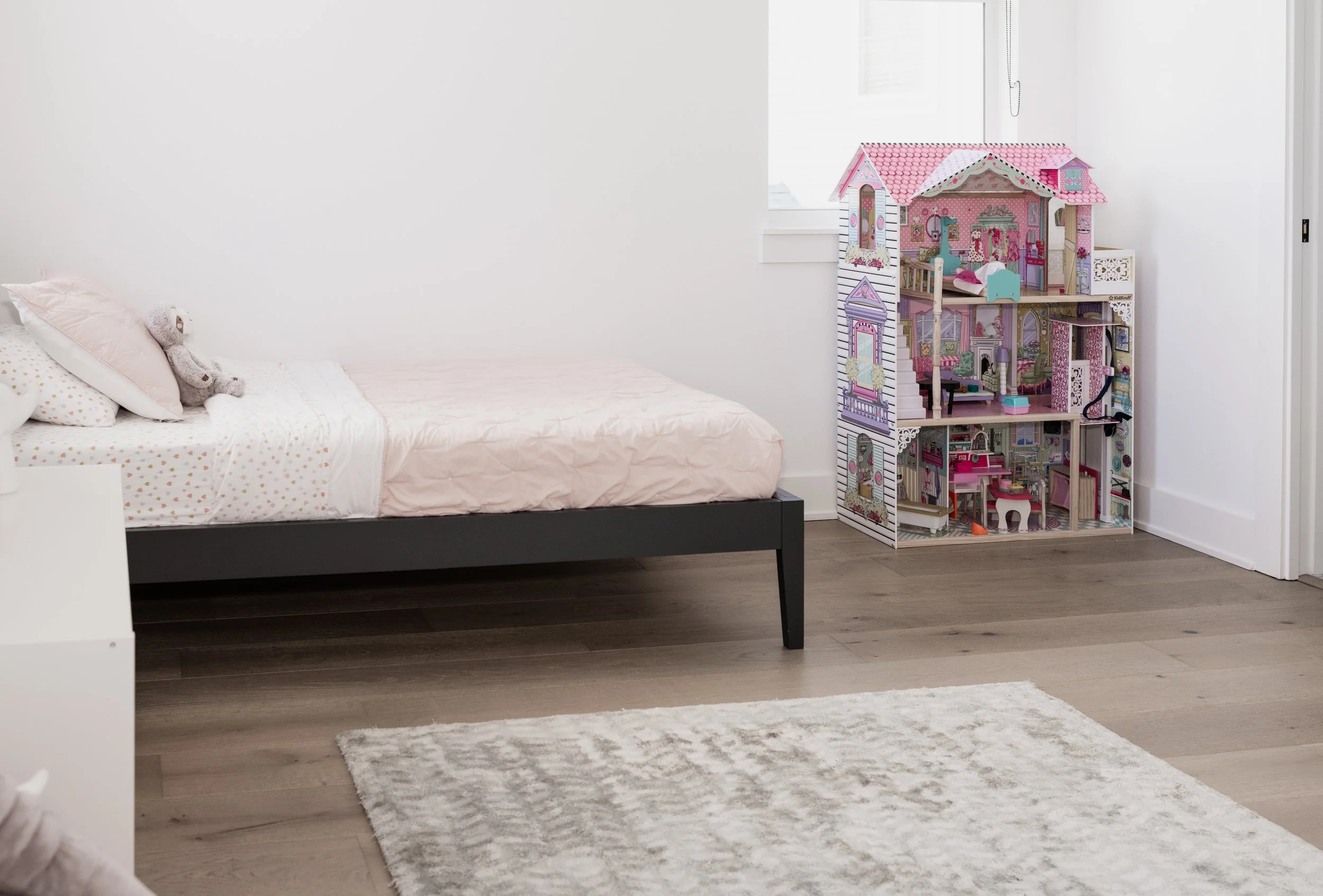 A bedroom with a bed on the left, pink bedding, and a gray teddy bear, and a large dollhouse with multiple rooms and pink accents in the corner near a window.