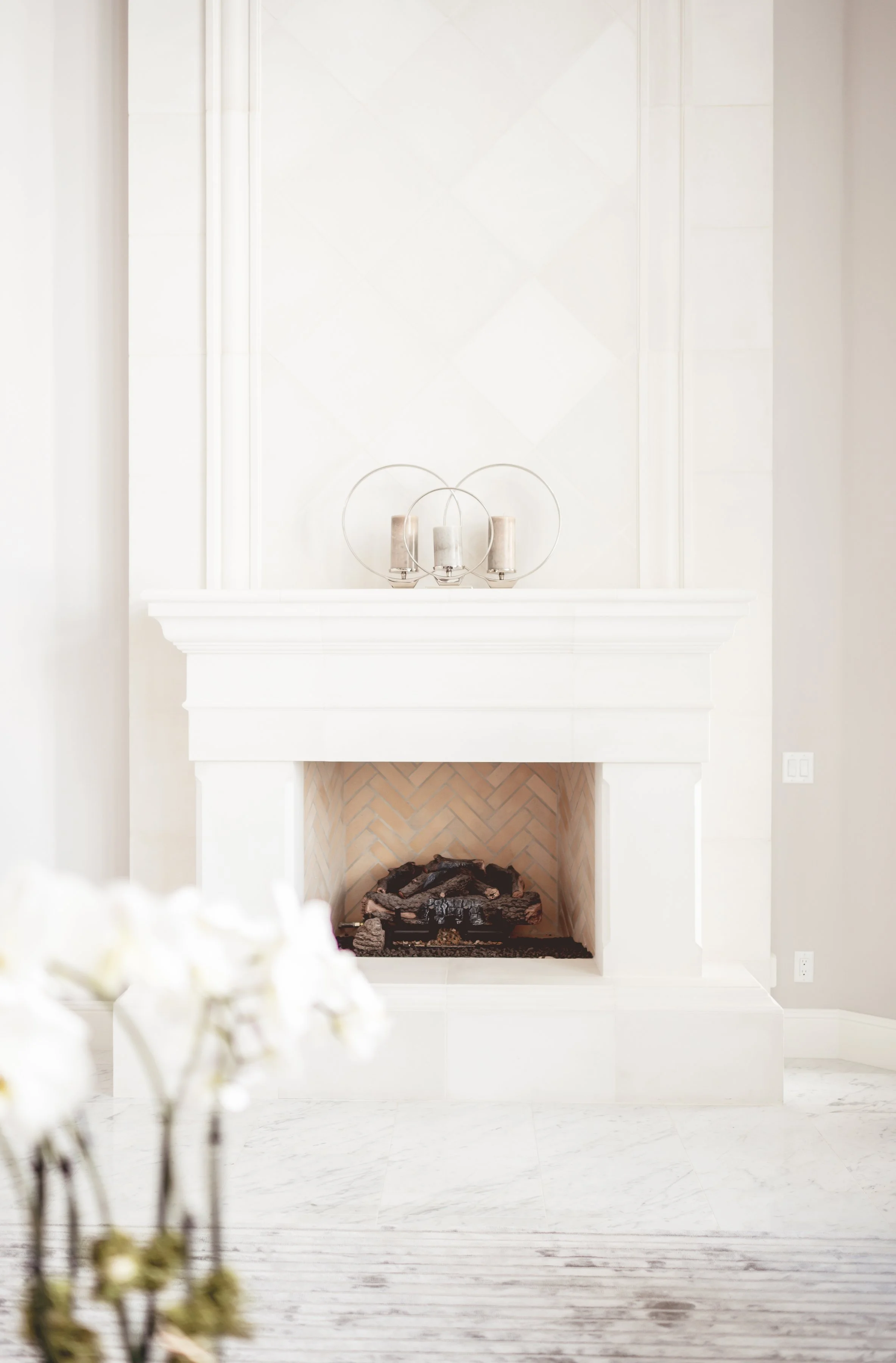 Minimalist fireplace with a white mantel and decorative candles on top, set against a white wall with light-colored flooring and a blurred floral arrangement in the foreground.