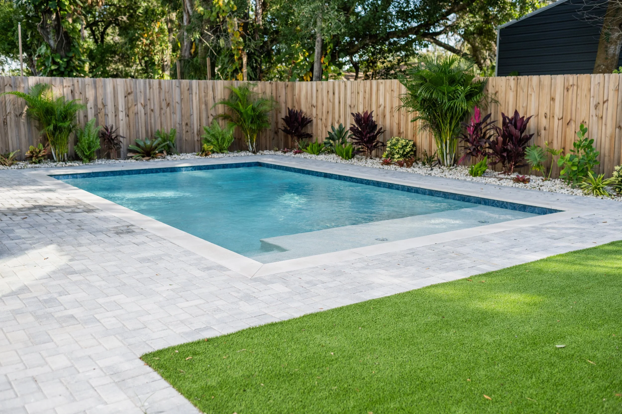 Backyard with a rectangular swimming pool, surrounded by gray stone pavers, green grass, and a wooden privacy fence with various plants and shrubs along the fence line.