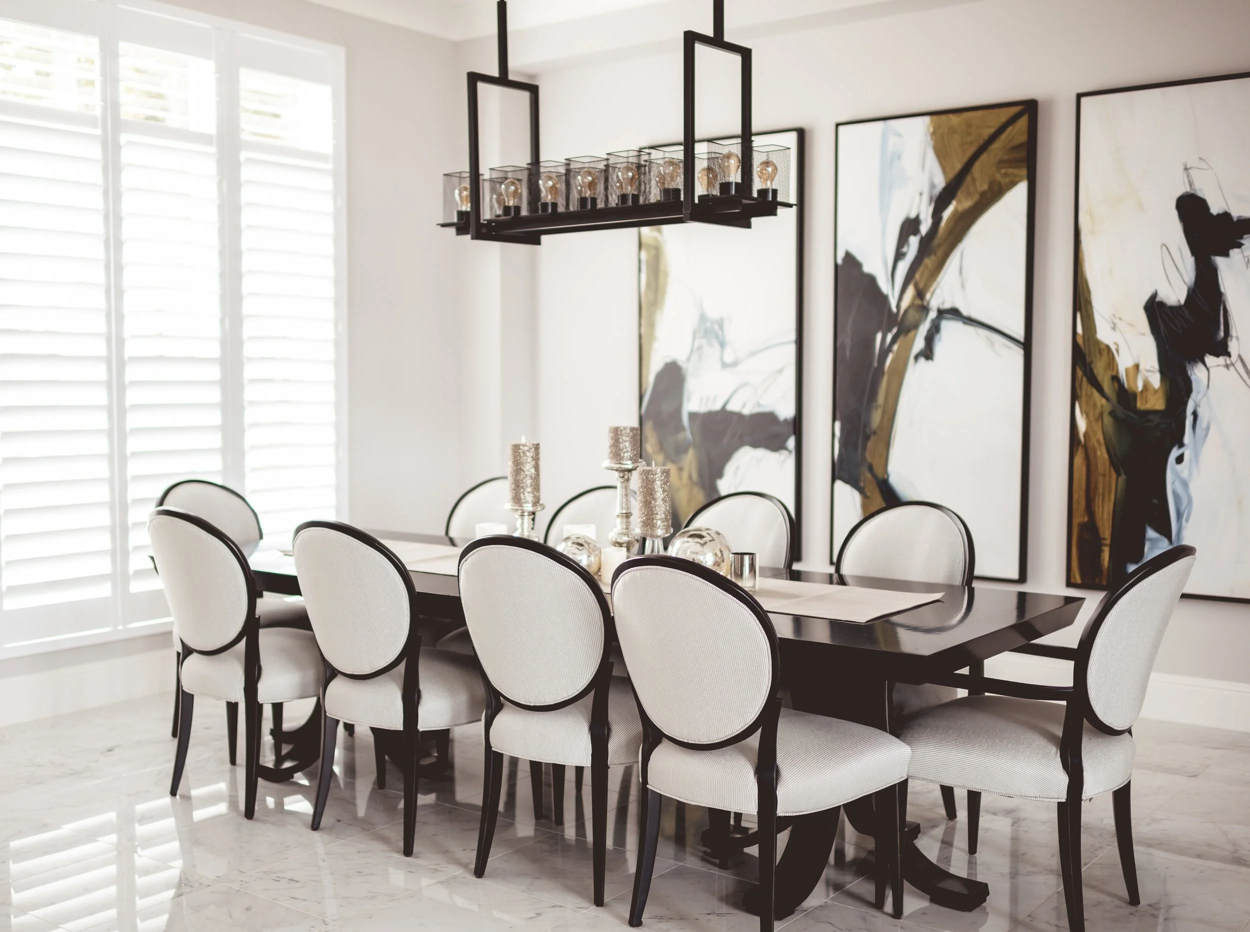 Modern dining room with a black table, white upholstered chairs with black frames, abstract wall art, and a black geometric chandelier, illuminated by natural light from plantation shutters.
