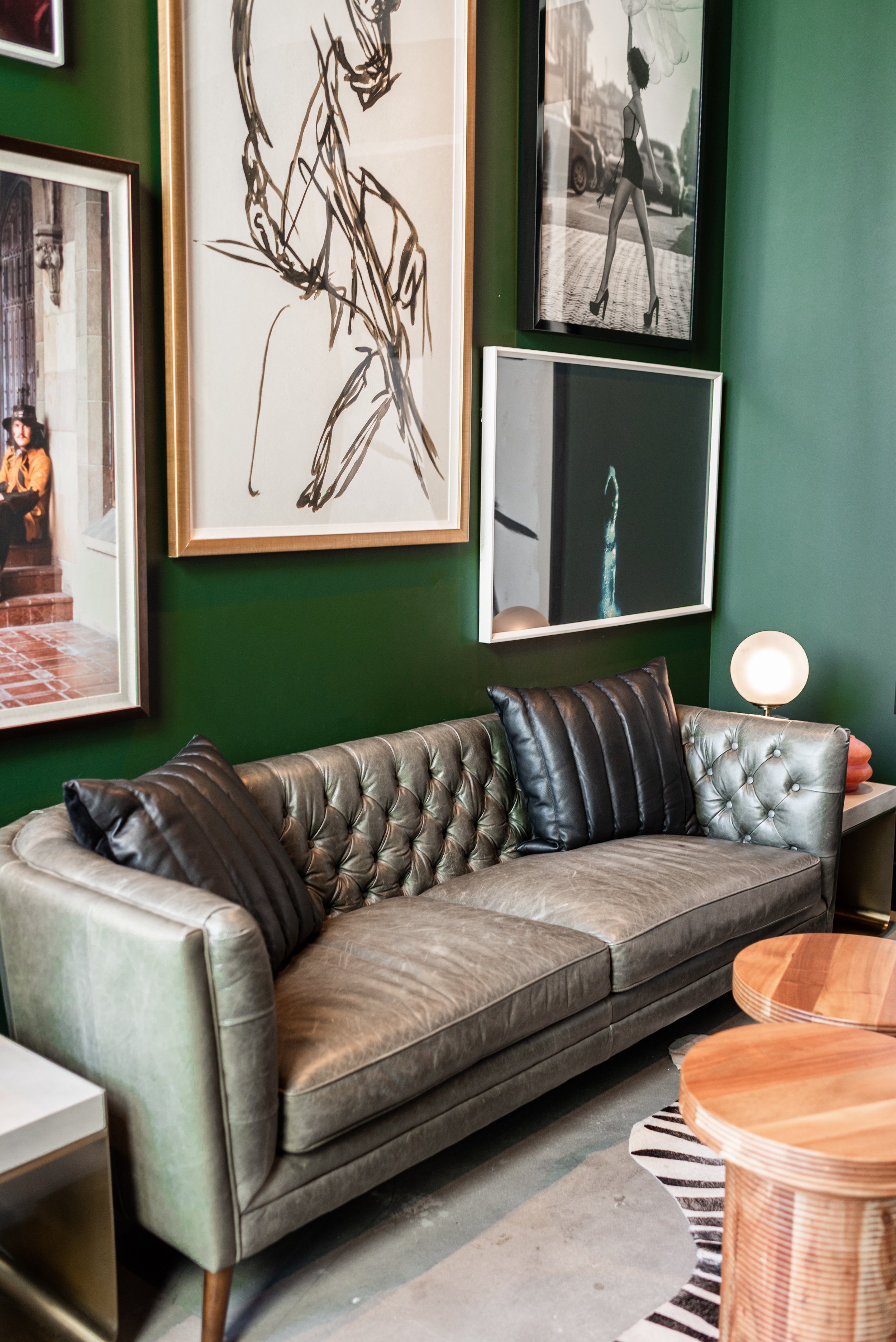 A living room with a gray tufted leather couch, black pillows, framed art on a green wall, including line sketches and photographs, a small white side table with a round lamp, and two wooden coffee tables.