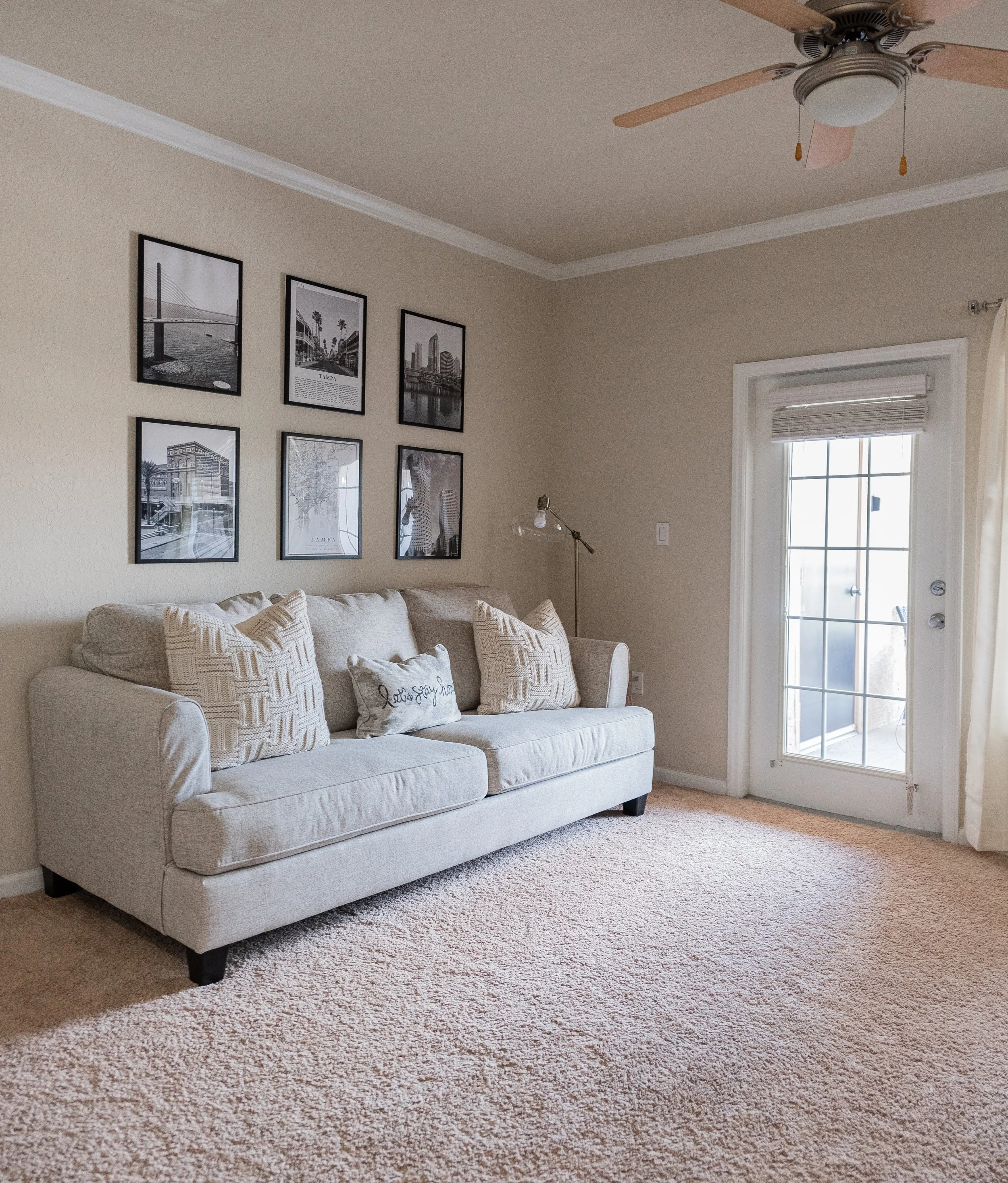 A living room with a beige sofa decorated with four patterned cushions and a small pillow that reads 'I love you'. Above the sofa is a wall with six framed black-and-white photographs. To the right of the sofa is a glass door with a curtain, leading 