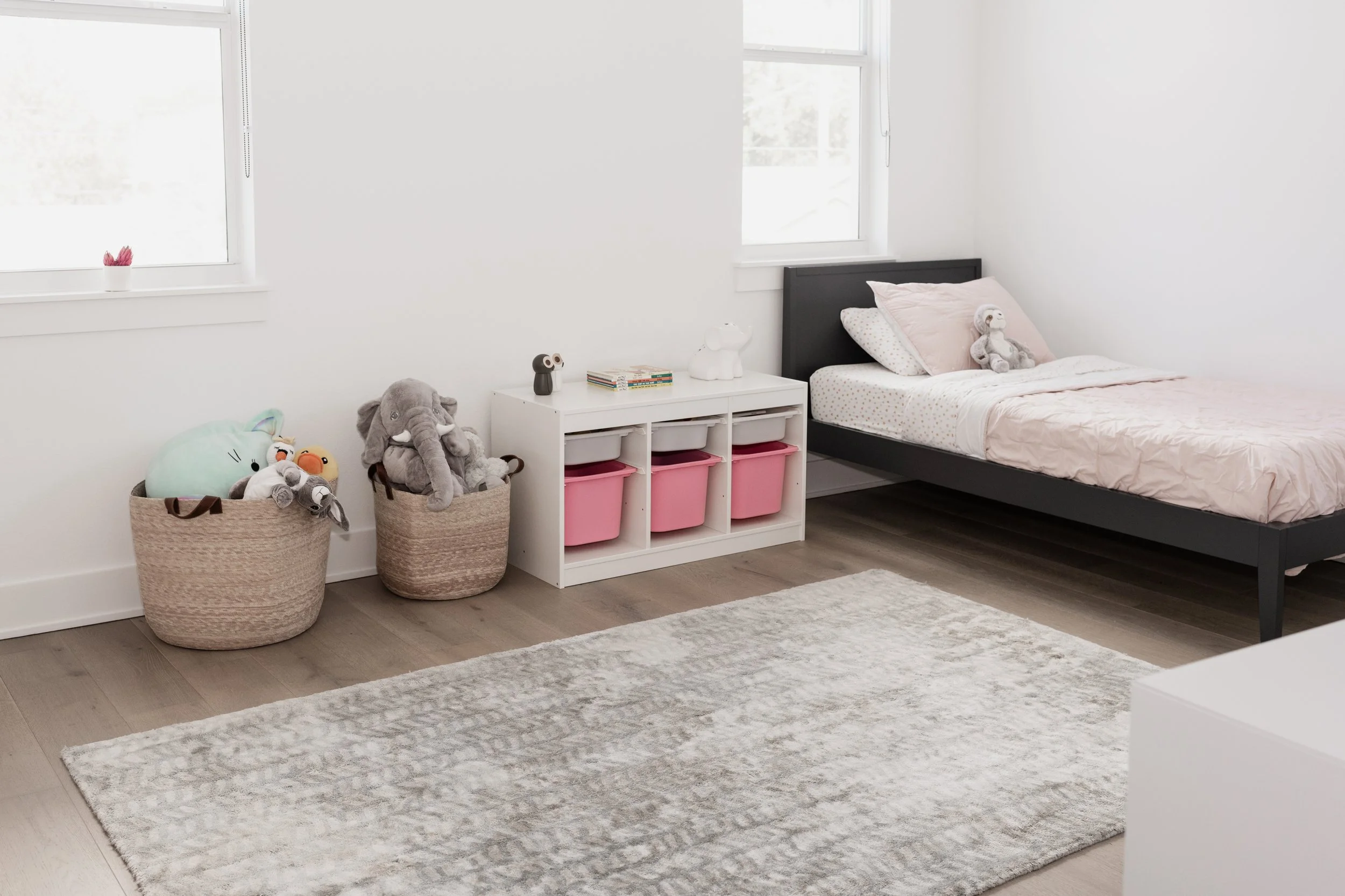 A child's bedroom with a bed, stuffed animals, pink storage bins, and a white window with sunlight.