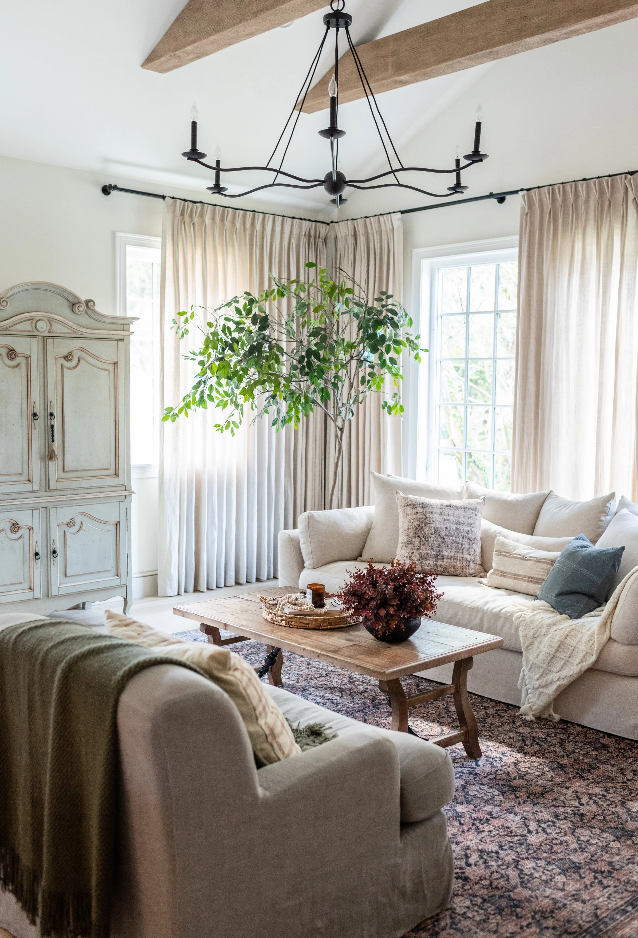 A cozy living room with a beige sectional sofa, a wooden coffee table with decorative items, a large potted plant, vintage-style cream-colored armoire, and four tall windows with light-colored curtains. A black chandelier hangs from the ceiling.