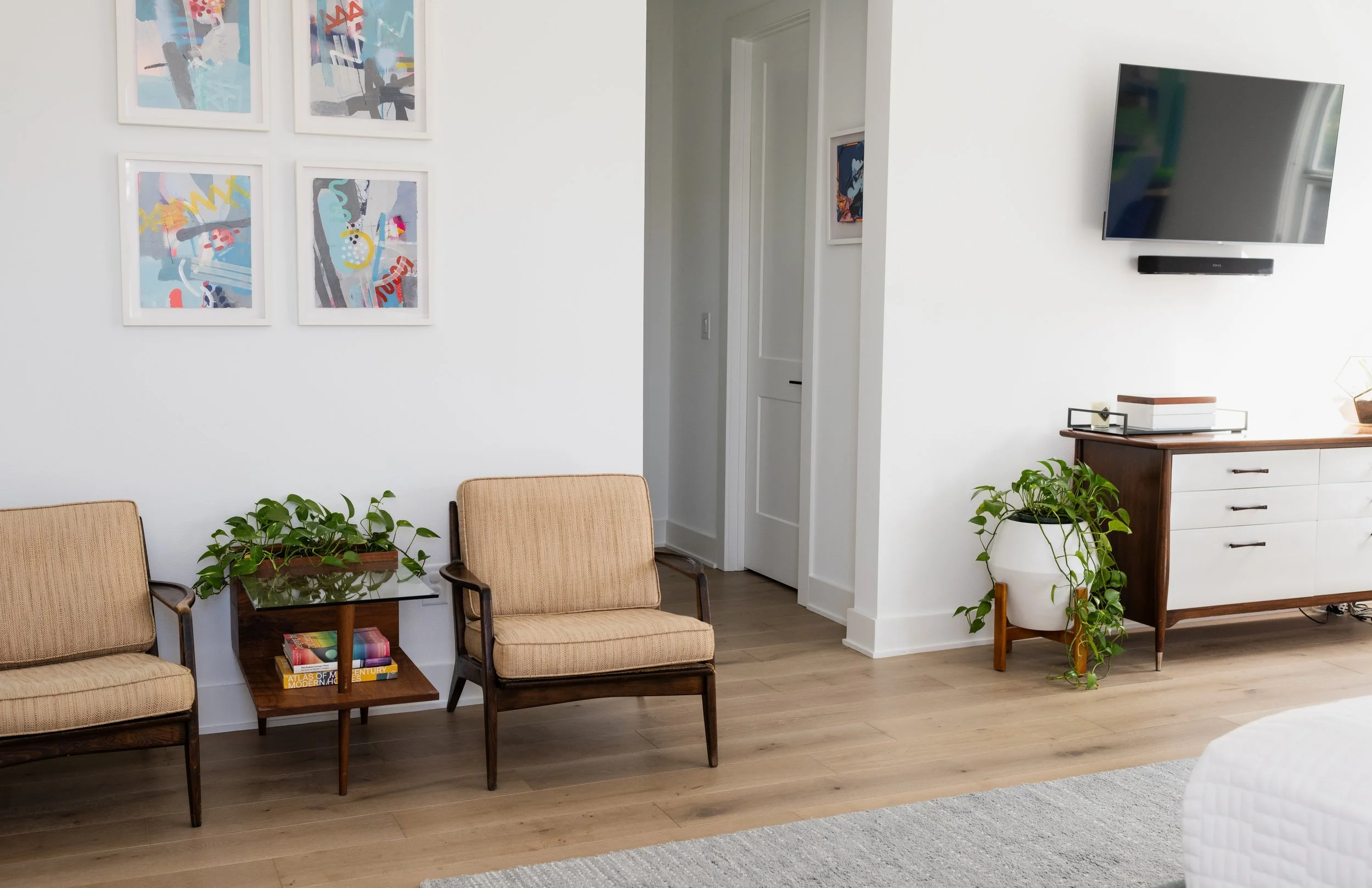 Living room with a wall-mounted TV, white walls, wooden furnishings, beige chairs, green plants, abstract art on the wall, and wooden flooring.
