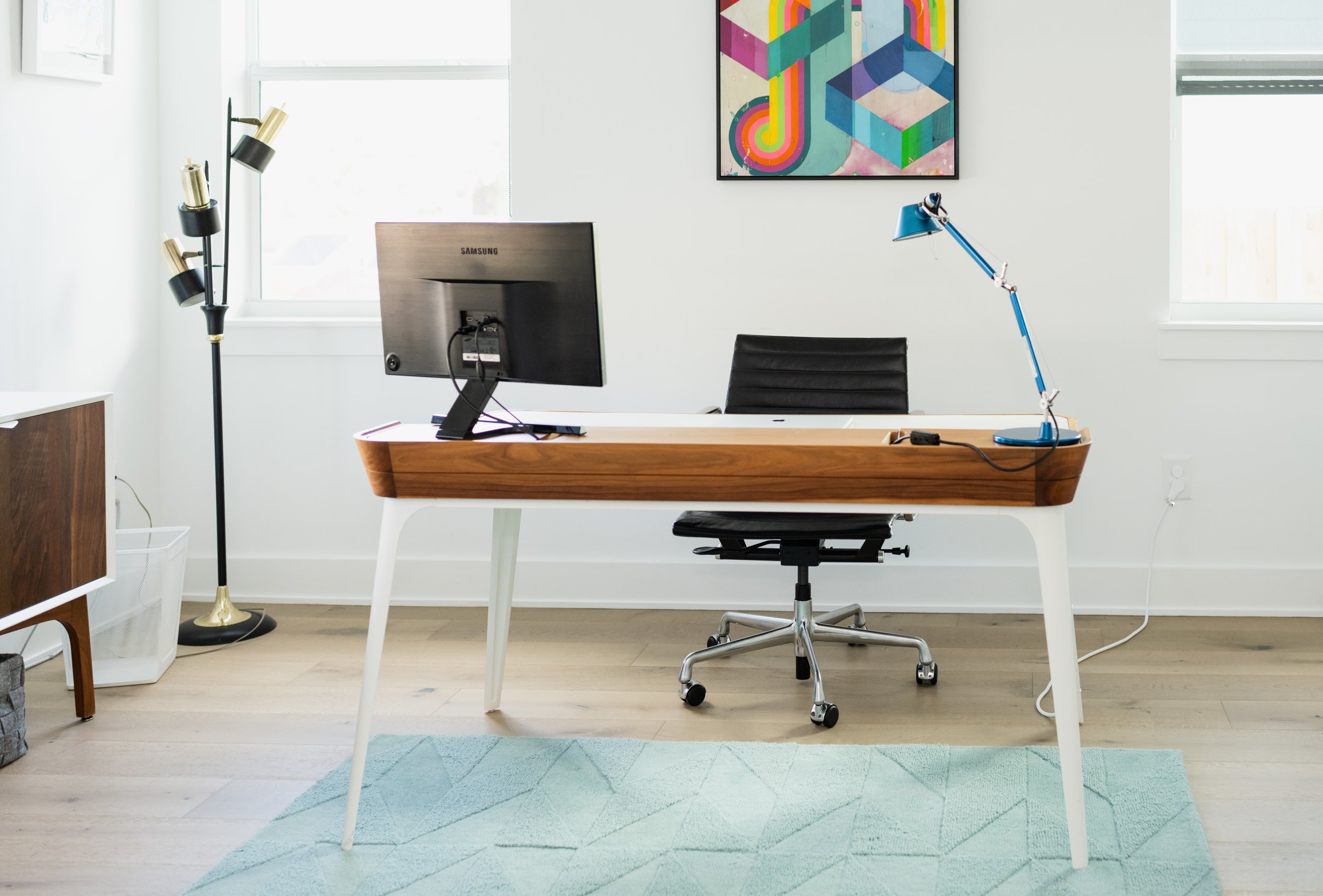 Modern home office with a wooden desk, a computer monitor, a black office chair, a blue desk lamp, and colorful artwork on a white wall.