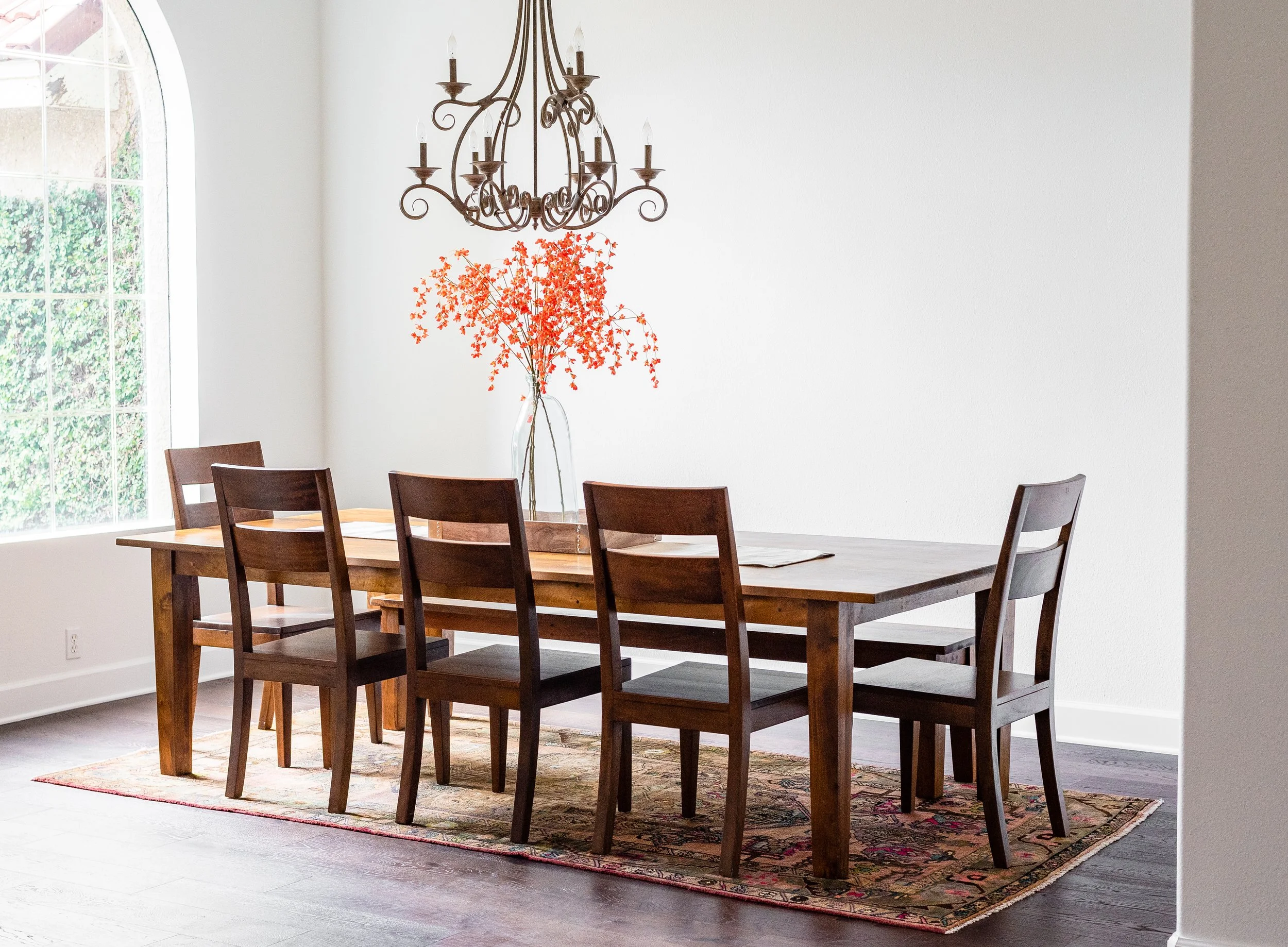 A dining room with a wooden table, six wooden chairs, a chandelier, a large vase with red-orange flowers, and a window with greenery outside.