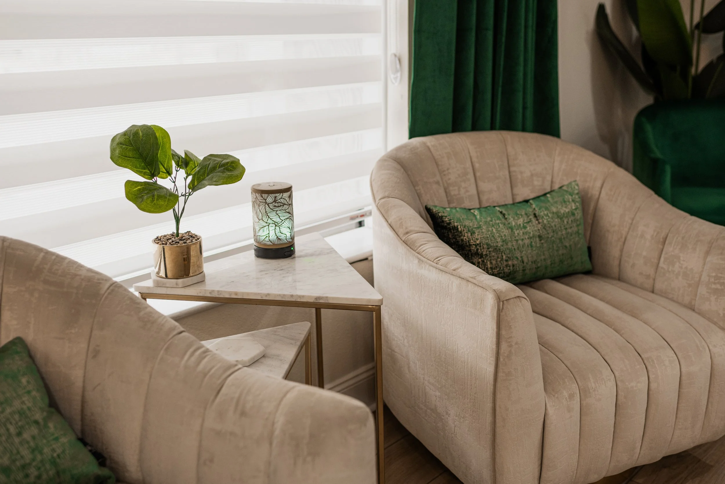 Living room with beige upholstered chairs, green pillows, a small marble side table with a potted plant, and a decorative lamp, next to white blinds and green curtains.