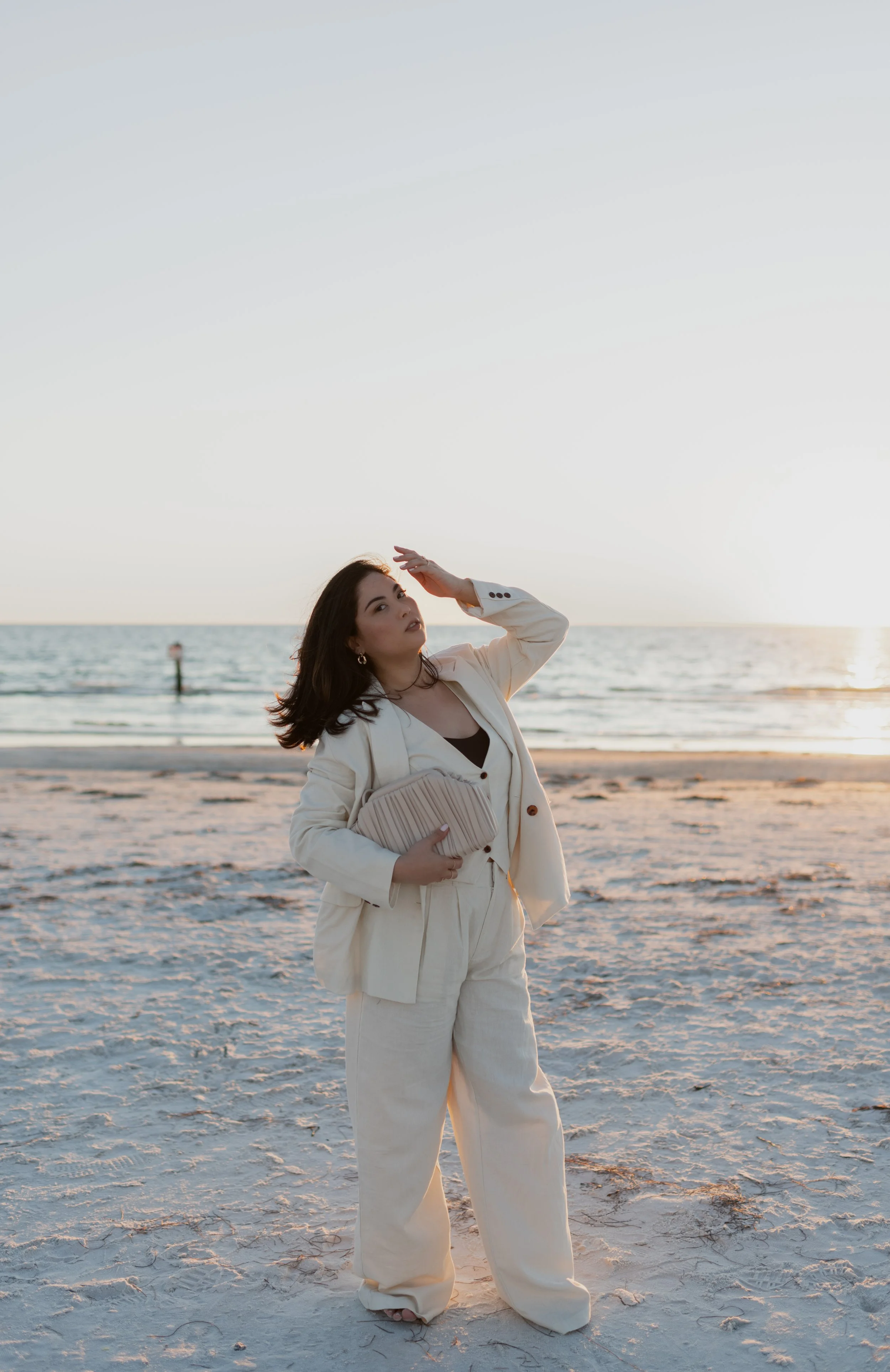A woman standing on a sandy beach during sunset, wearing a cream-colored suit and holding a pleated clutch, with the ocean in the background.