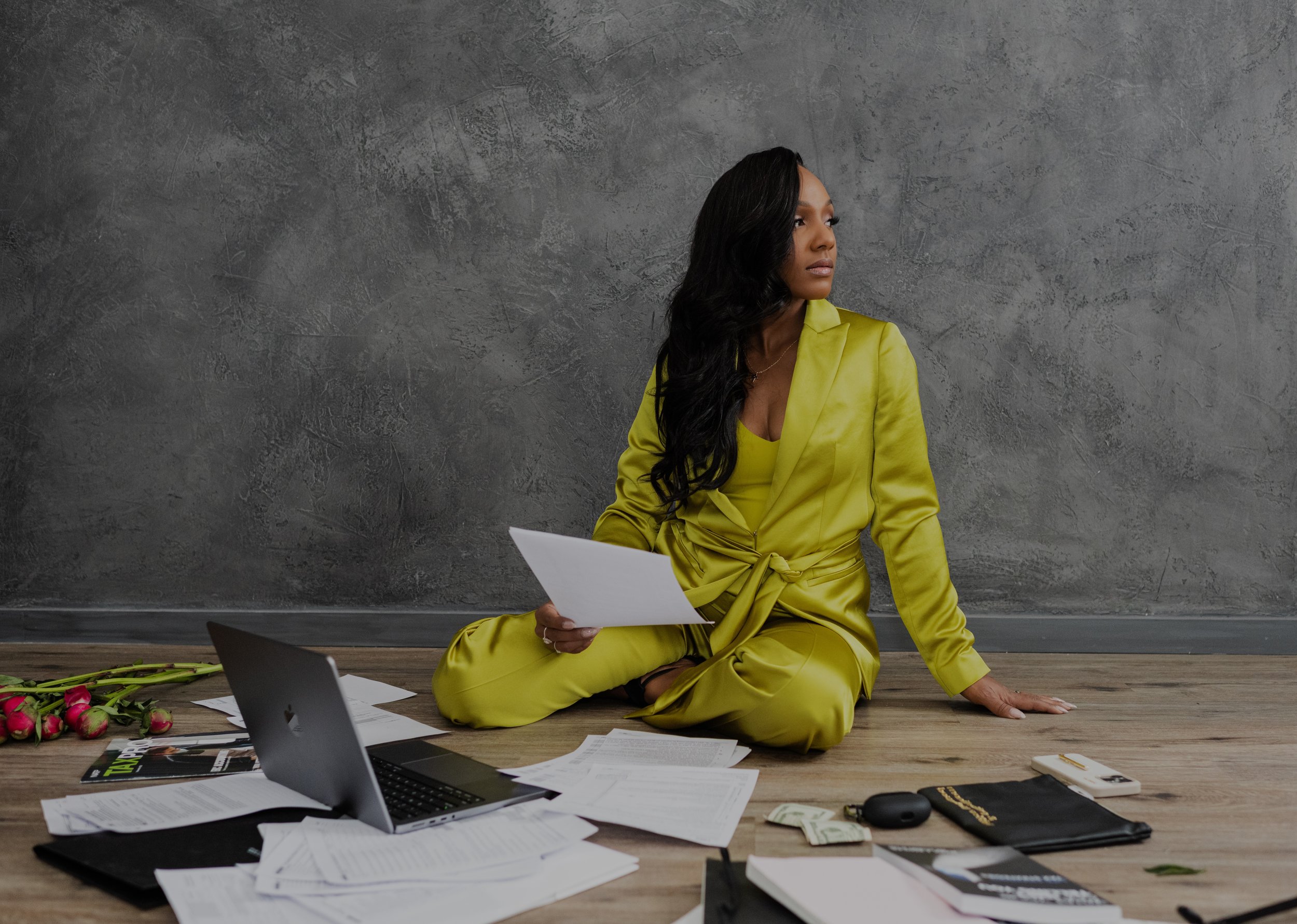 A woman in a yellow suit sitting on a wooden floor in front of a gray wall with scattered papers, a laptop, flowers, and office supplies around her, looking to the side.