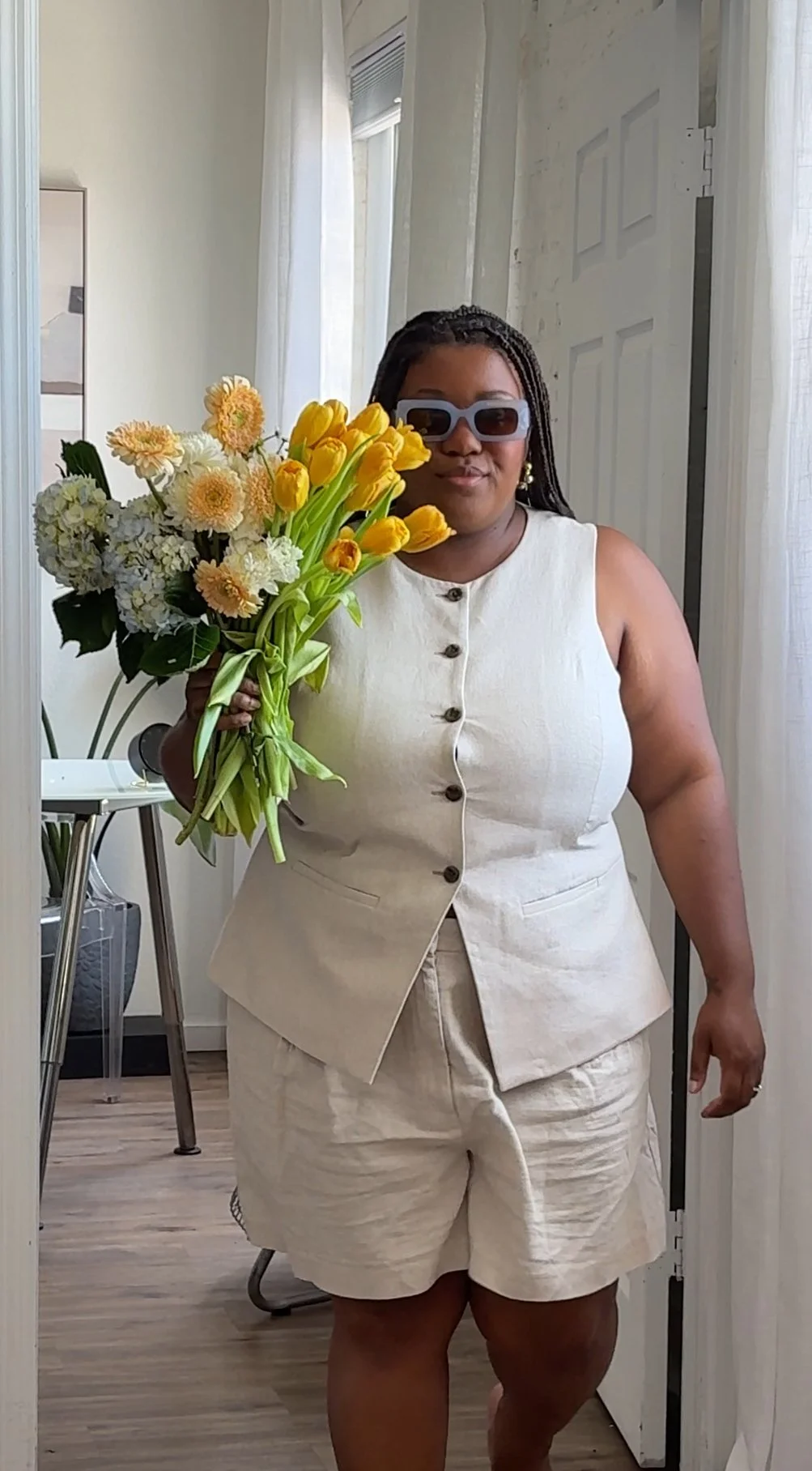 A woman in a beige sleeveless vest and shorts holding a bouquet of yellow and white flowers, wearing sunglasses, standing inside a room with white curtains and a door.