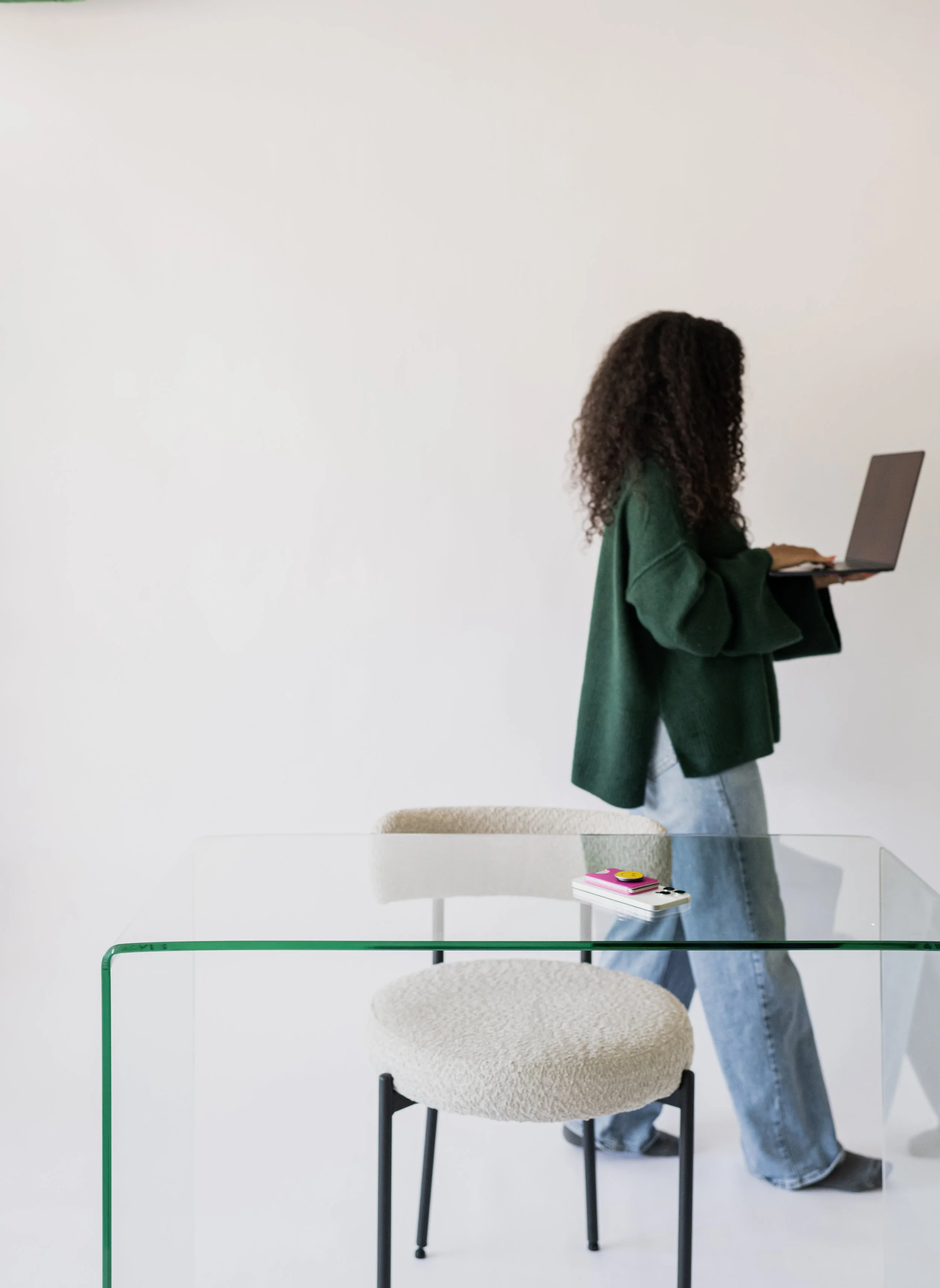 A woman with curly hair wearing a green coat and light blue jeans, working on a laptop in a minimalistic room with white walls and furniture.