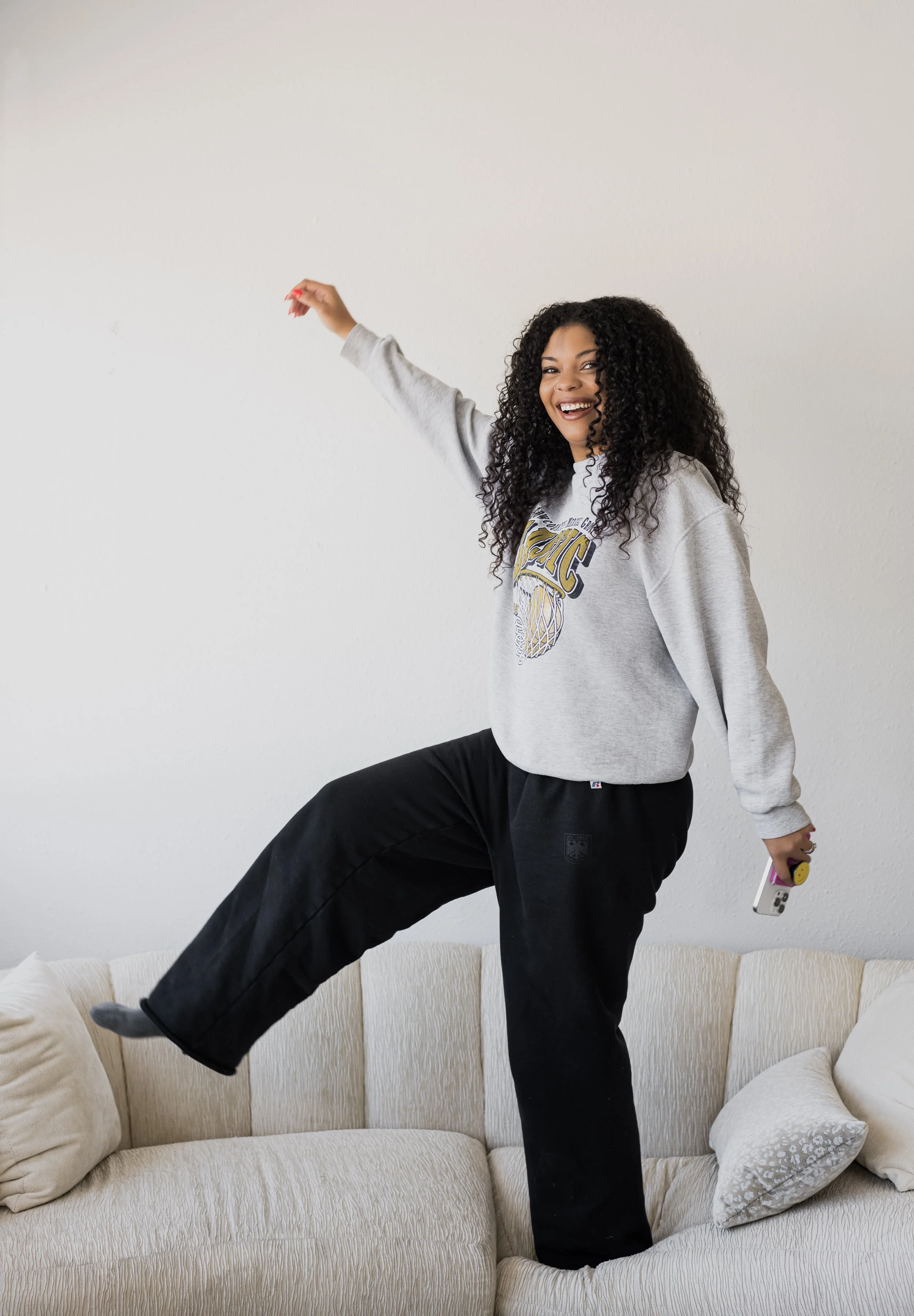 A woman with curly hair standing on a cream-colored sofa, smiling, with one foot raised, holding a phone in her right hand, in a casual indoor setting.