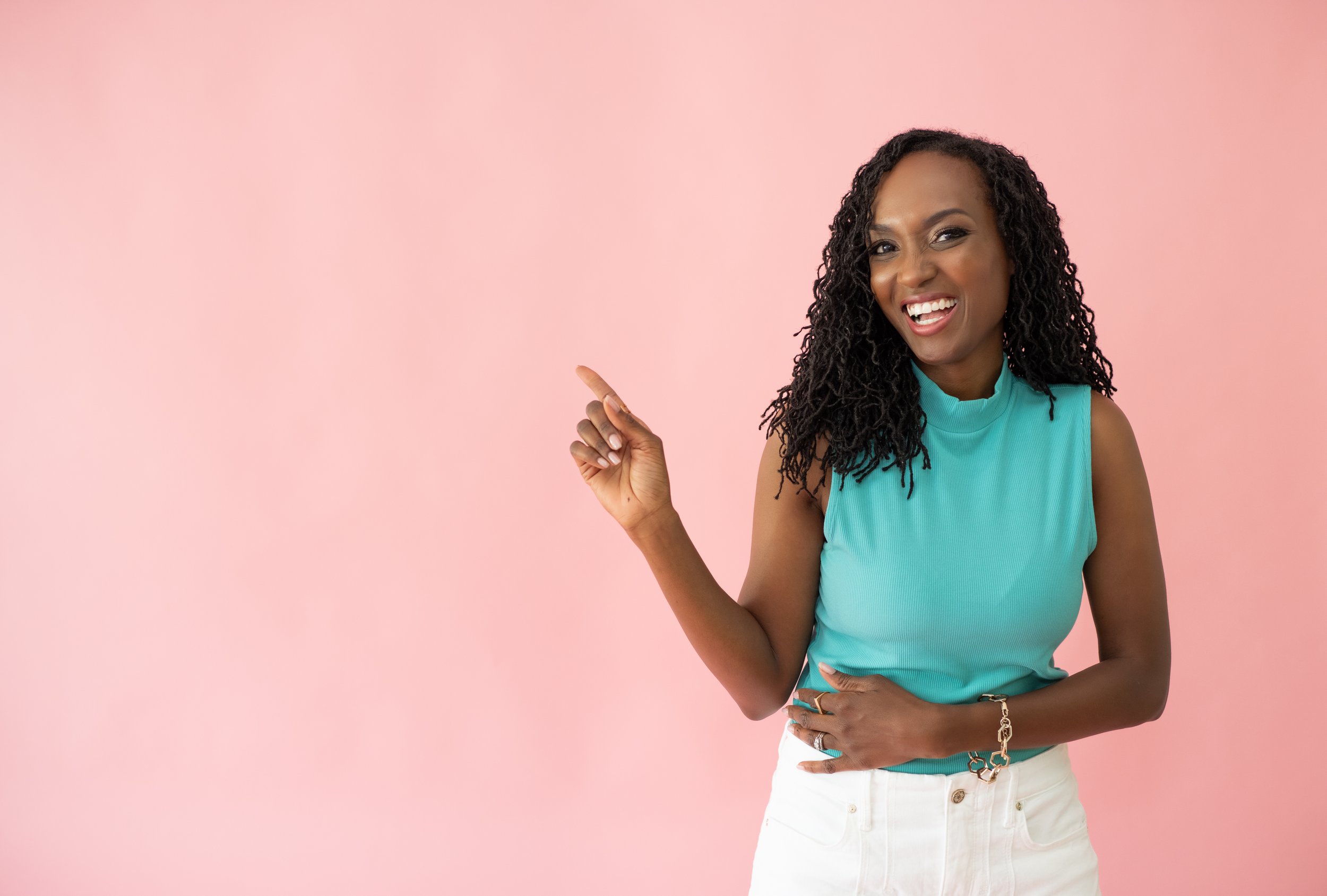 A woman with dark curly hair, wearing a sleeveless turquoise top and white pants, smiling and pointing to her left against a pink background.