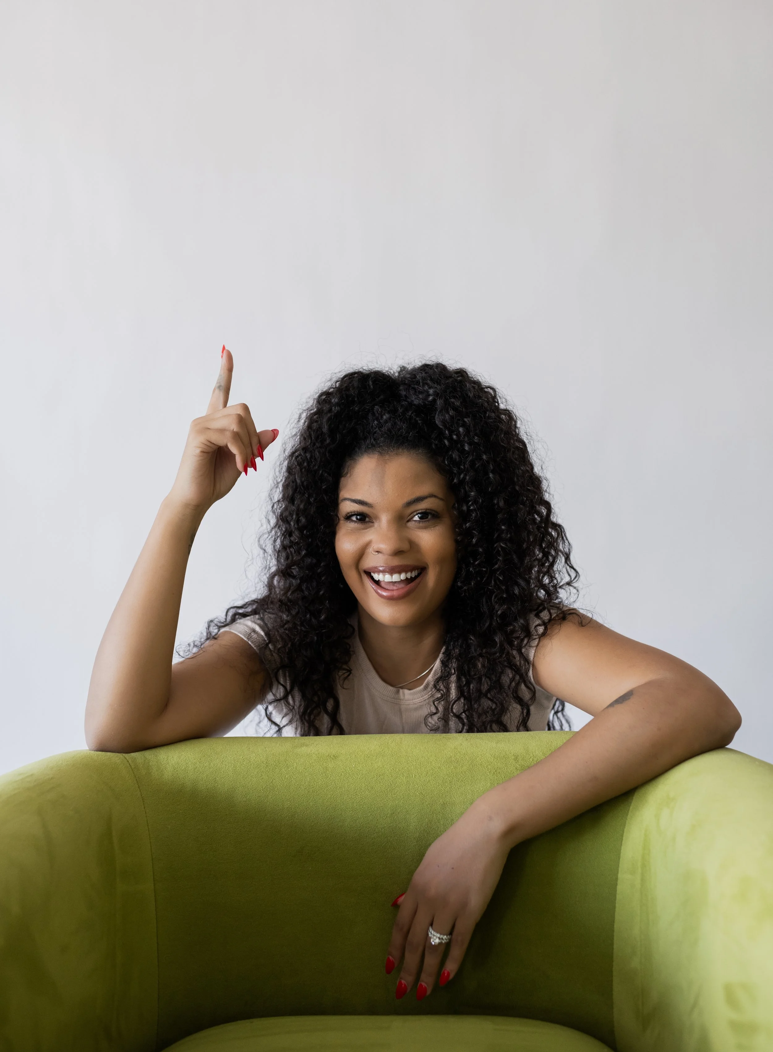 A woman with curly hair smiling and pointing upwards while leaning on a green chair against a plain white wall.