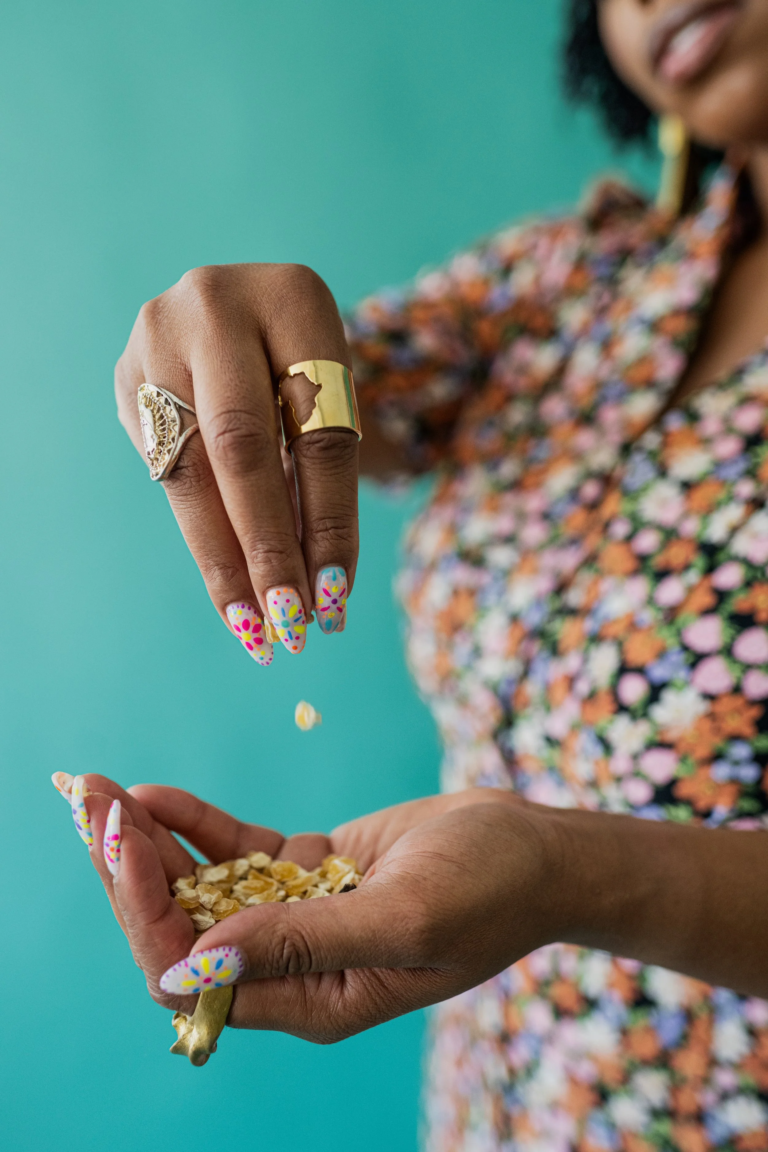 A woman with dark skin showing her hands, wearing colorful rings and decorated nail polish, pouring cereal from one hand into the other against a solid teal background.