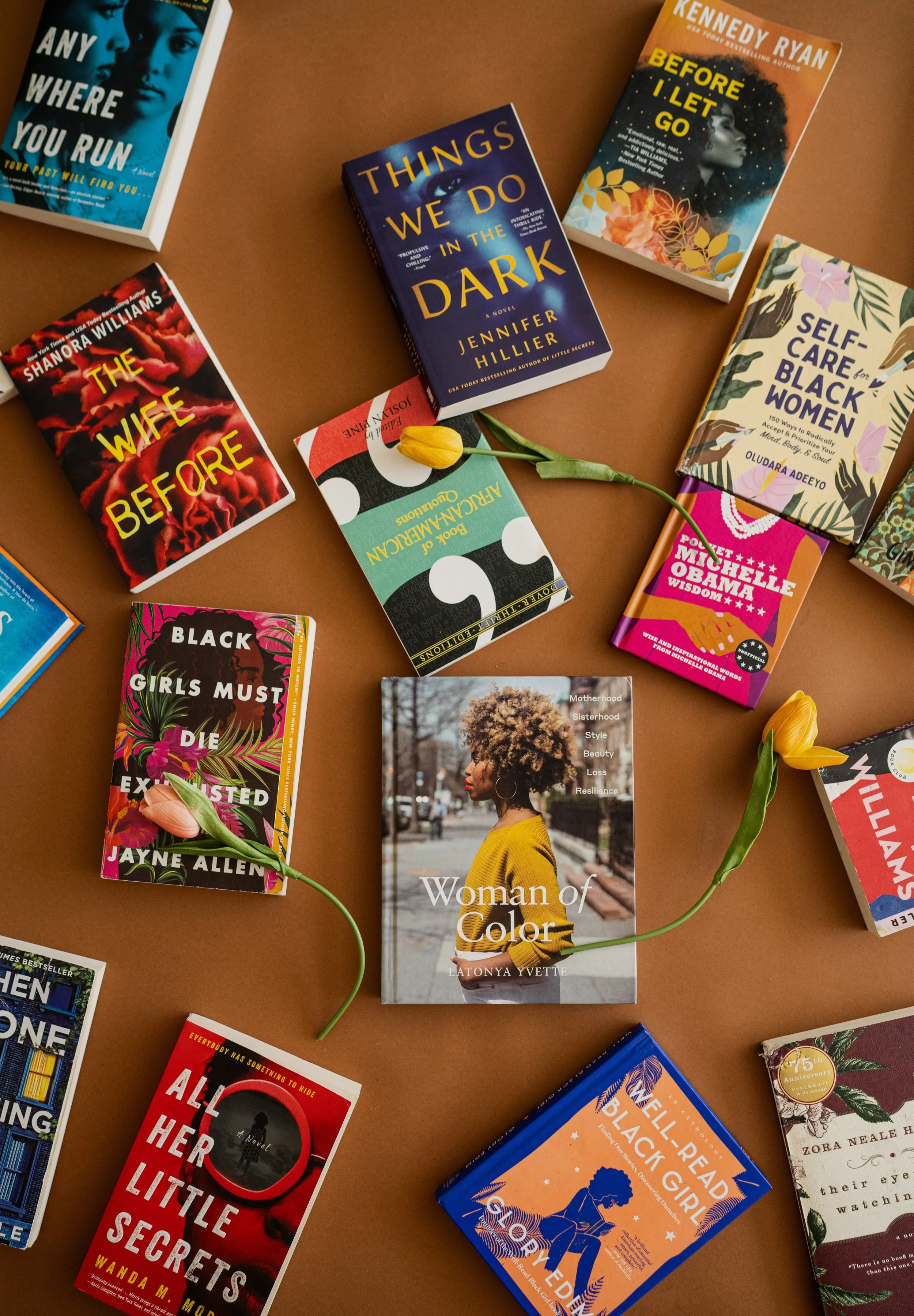 Collection of books on a brown surface with two yellow tulips intertwined among them, including 'Woman of Color' by Latonya Yvette, 'The Wife Before' by Shannon Williams, 'Things We Do in the Dark' by Jennifer Hillier, 'Before I Let Go' by Kennedy Ry