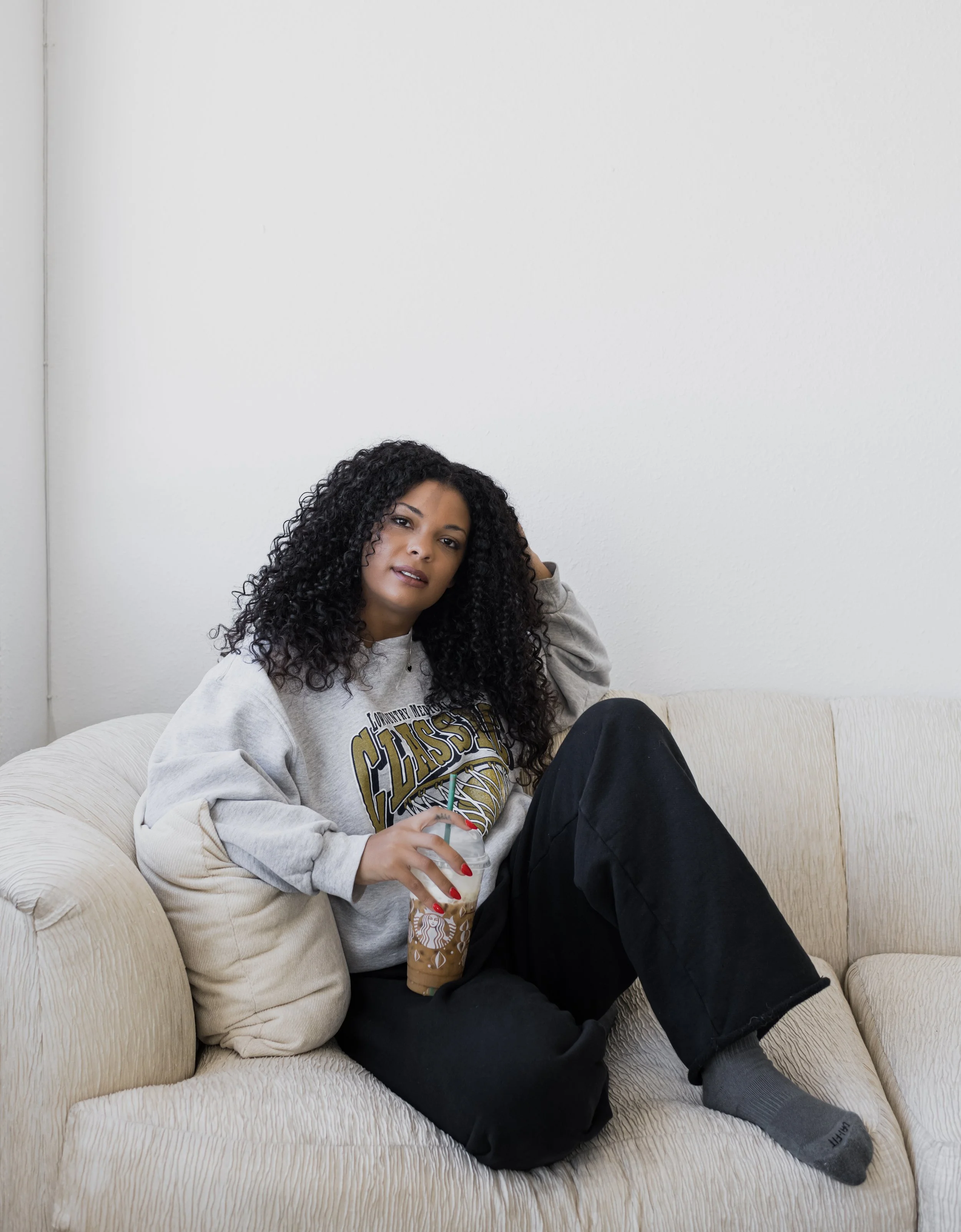 Young woman with curly hair sitting on a beige couch, holding a Starbucks iced coffee drink, wearing a gray sweatshirt and black pants, with one leg crossed and relaxed posture.