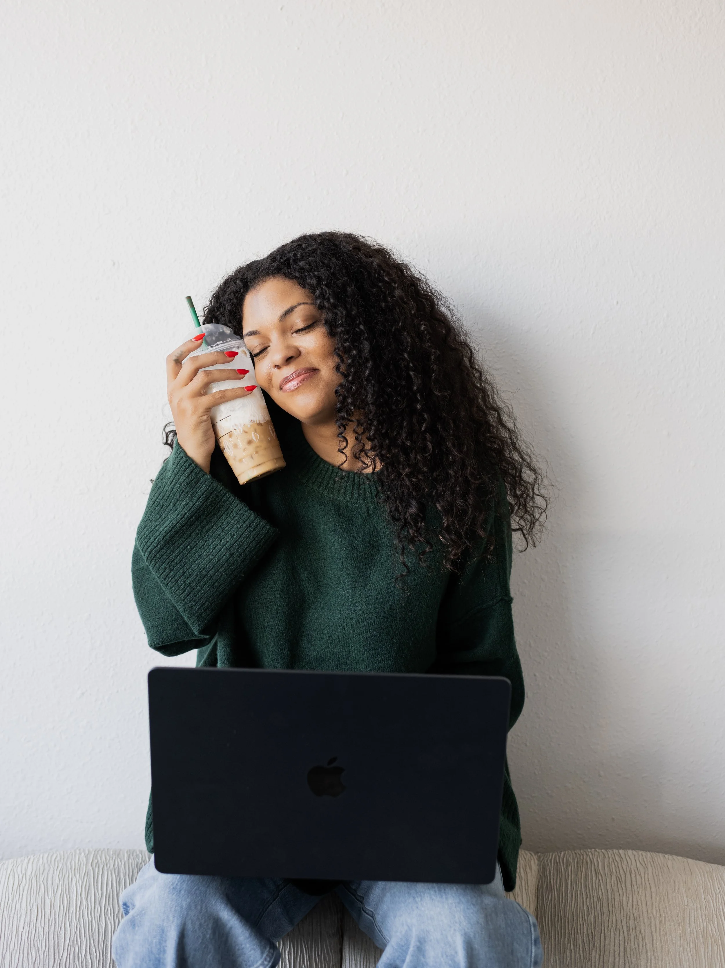 A woman with curly hair sitting on a beige sofa with a black laptop on her lap, holding a Starbucks frappuccino with whipped cream, smiling with her eyes closed, wearing a green sweater and light blue jeans, against a white wall.