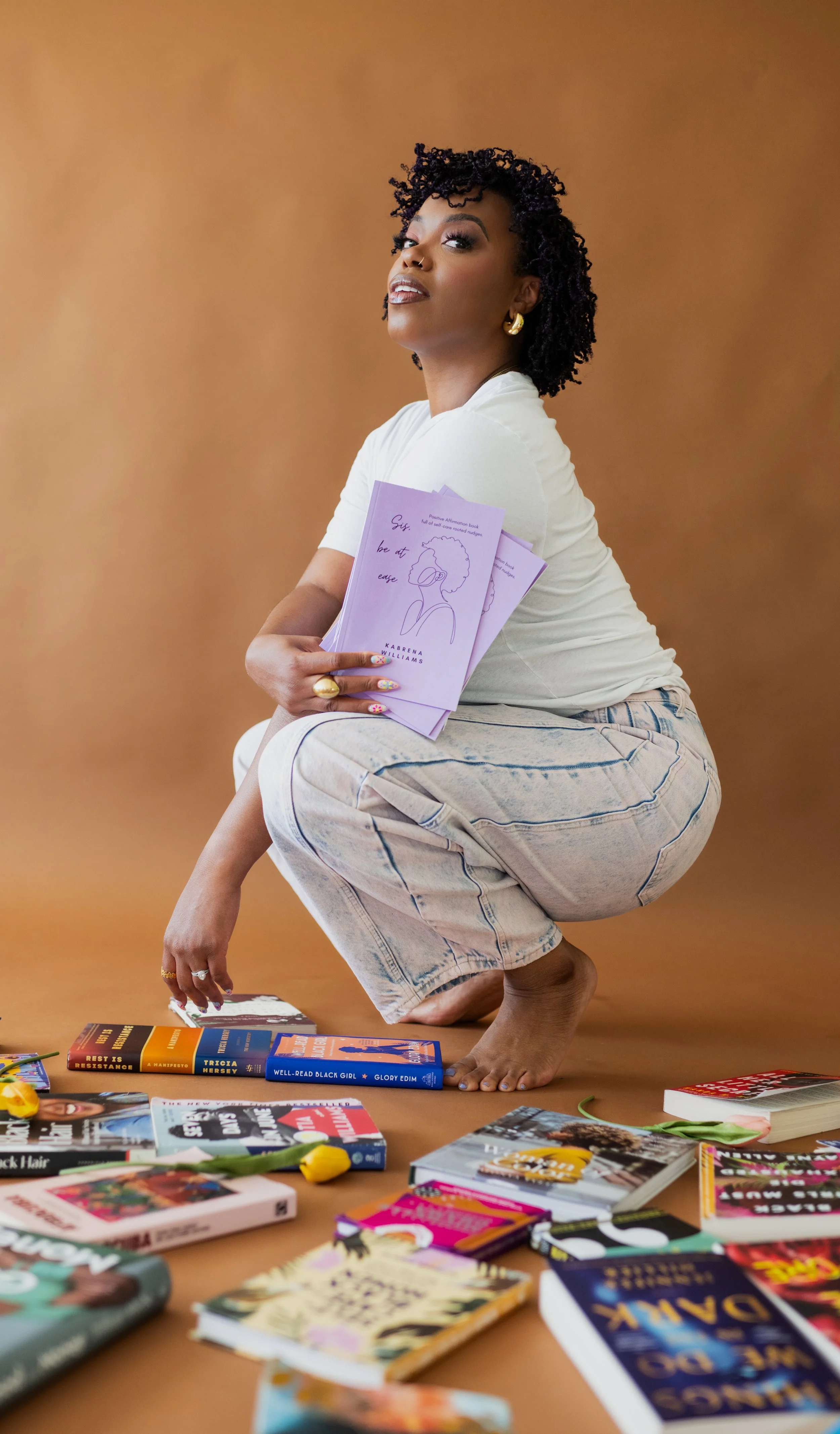 A woman squatting barefoot on a hardwood floor, holding purple books with a drawn face on the cover, surrounded by colorful books and yellow flowers on the floor, wearing a white t-shirt and light-colored jeans.