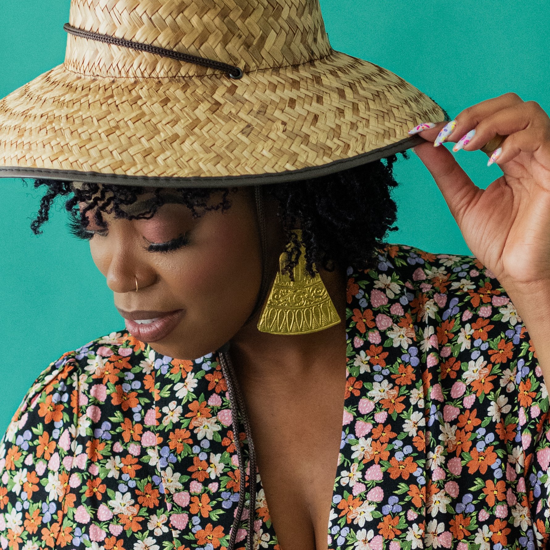 Woman wearing a large straw hat, floral patterned top, gold earrings, and a nose ring, with a teal background.