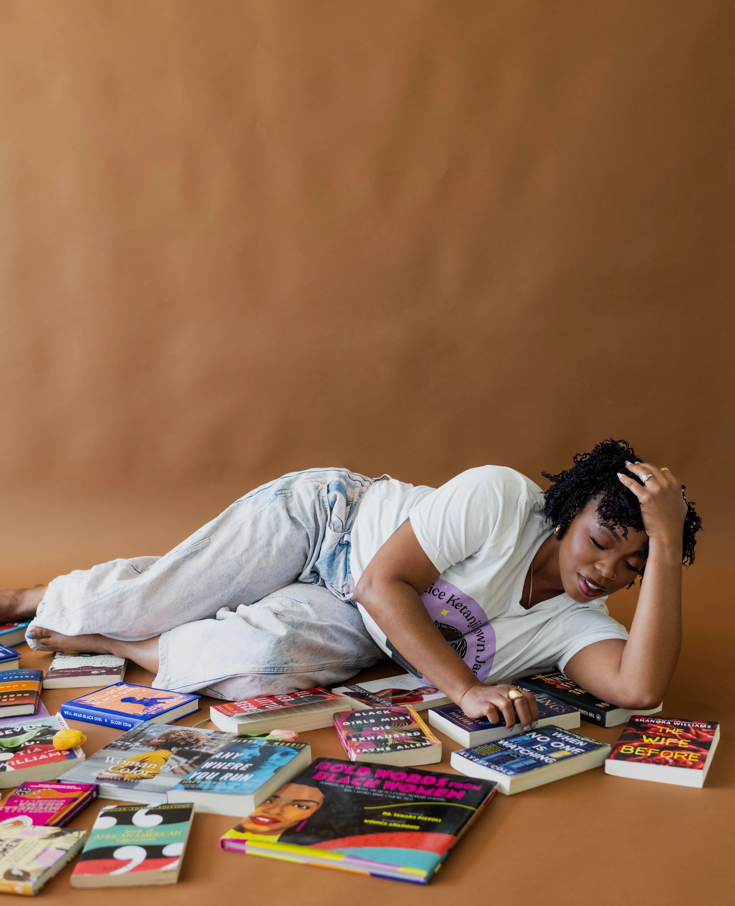 A woman lying on the floor surrounded by a variety of books, touching her forehead with one hand, appearing to be overwhelmed or stressed.
