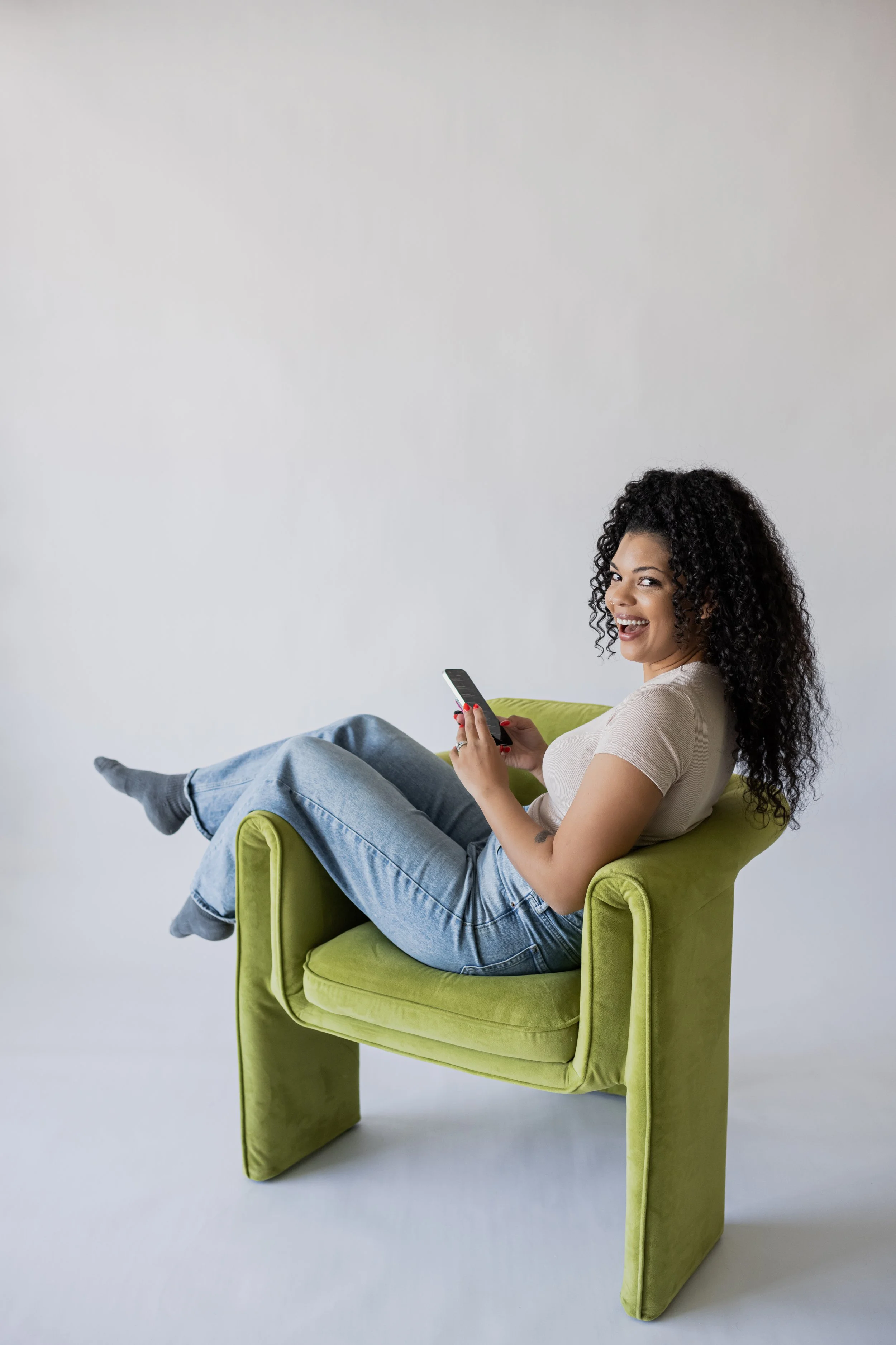 A woman with curly hair smiling and sitting casually on a lime green armchair while holding a smartphone