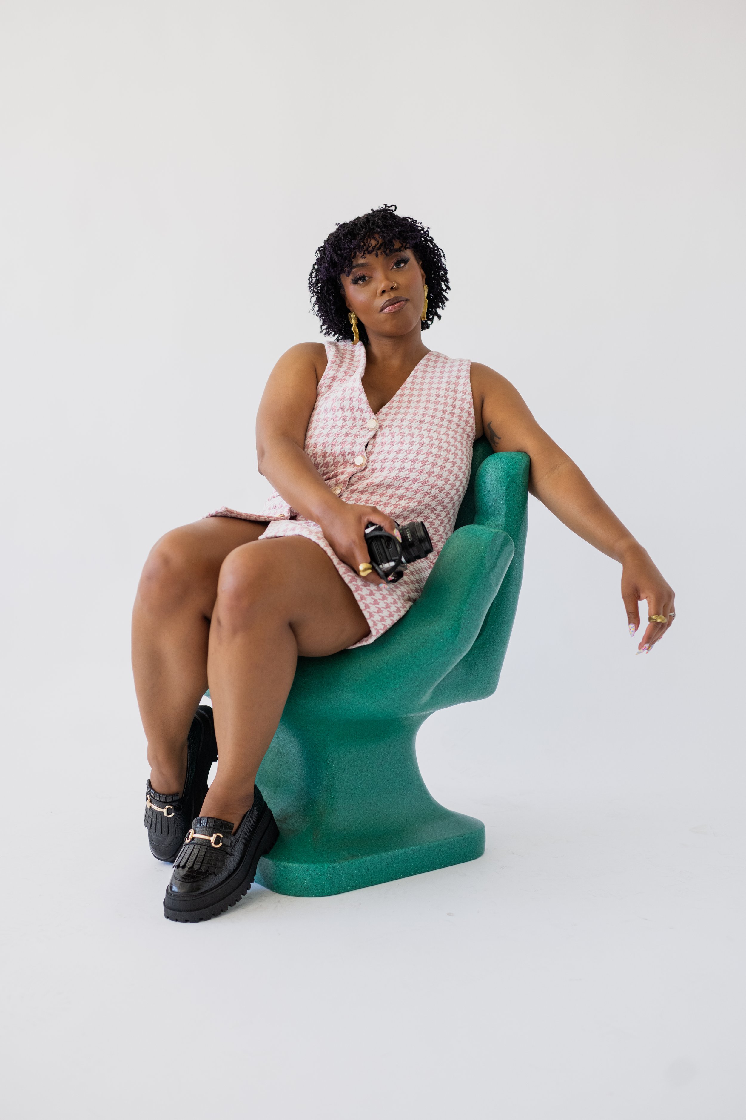 A woman with curly black hair sitting on a green modern chair, wearing a pink and white patterned sleeveless outfit, holding a camera, and wearing black shoes, in a minimal white studio background.