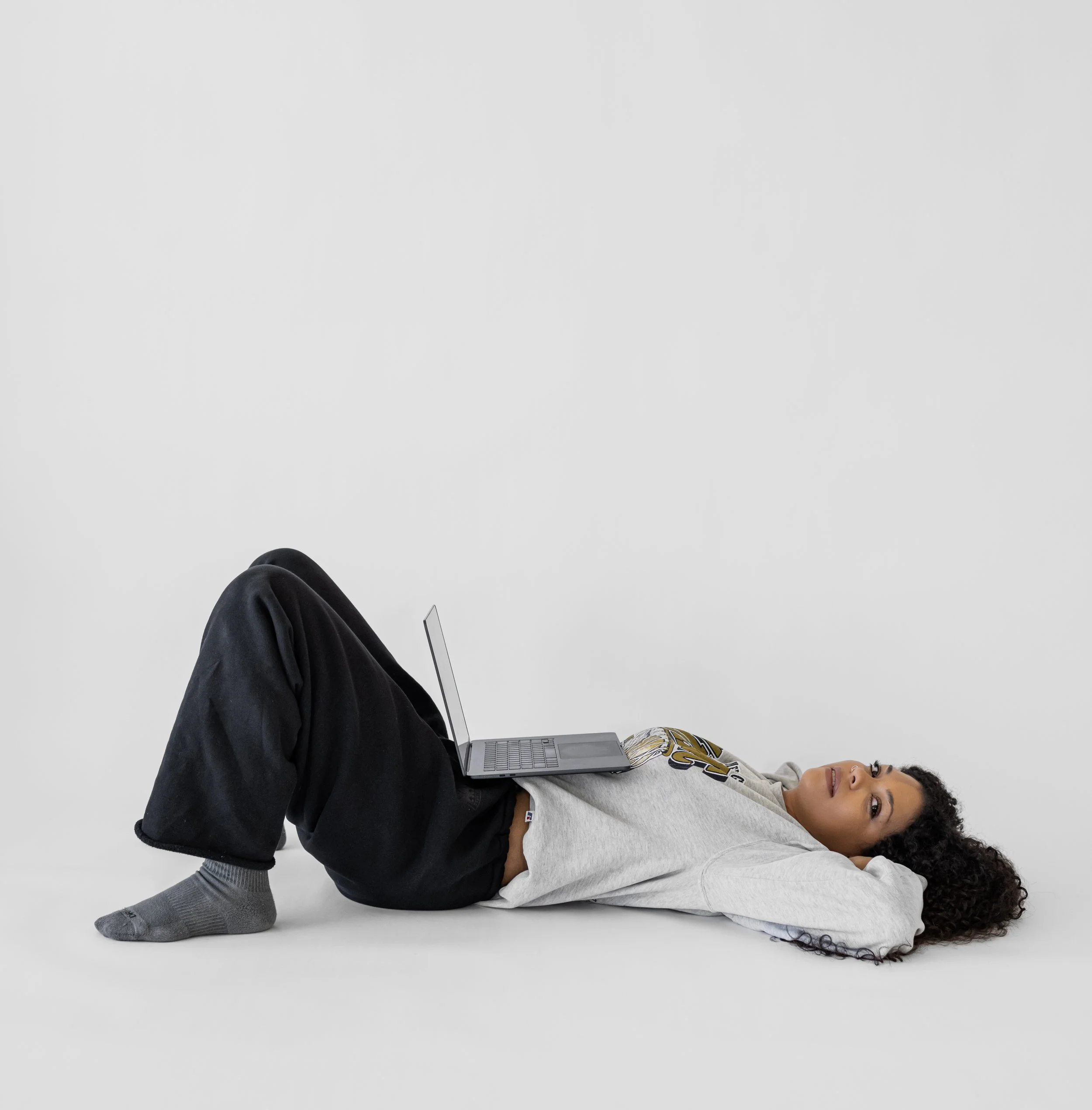 Young woman lying on her back on a plain white background with a laptop resting on her stomach, wearing a gray hoodie, black sweatpants, and gray socks, looking up at the camera.