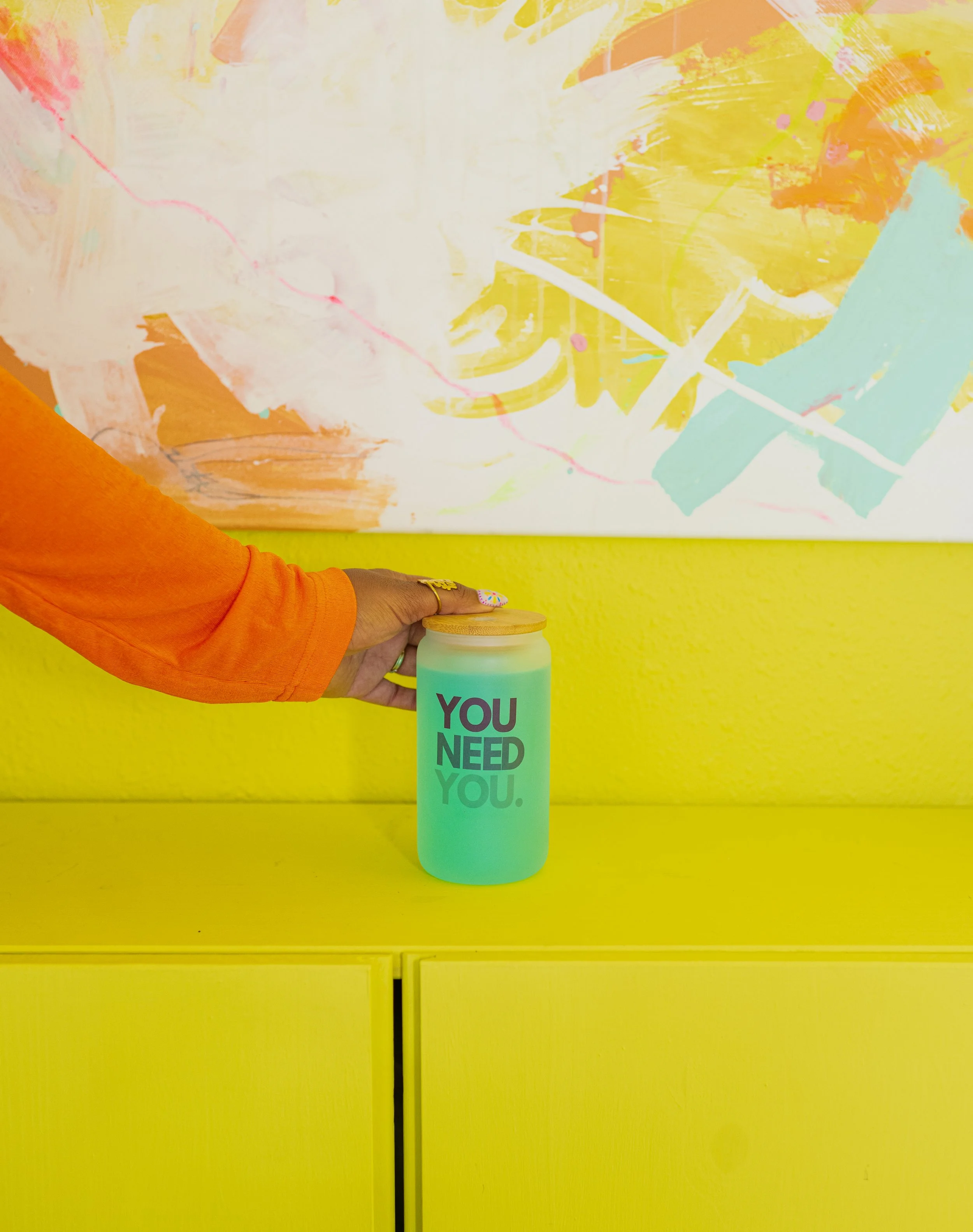 Person in an orange sleeve holding a light green jar with a wooden lid that has the words "YOU NEED YOU." written on it. The jar is placed on a bright yellow surface, with an abstract colorful painting in the background.