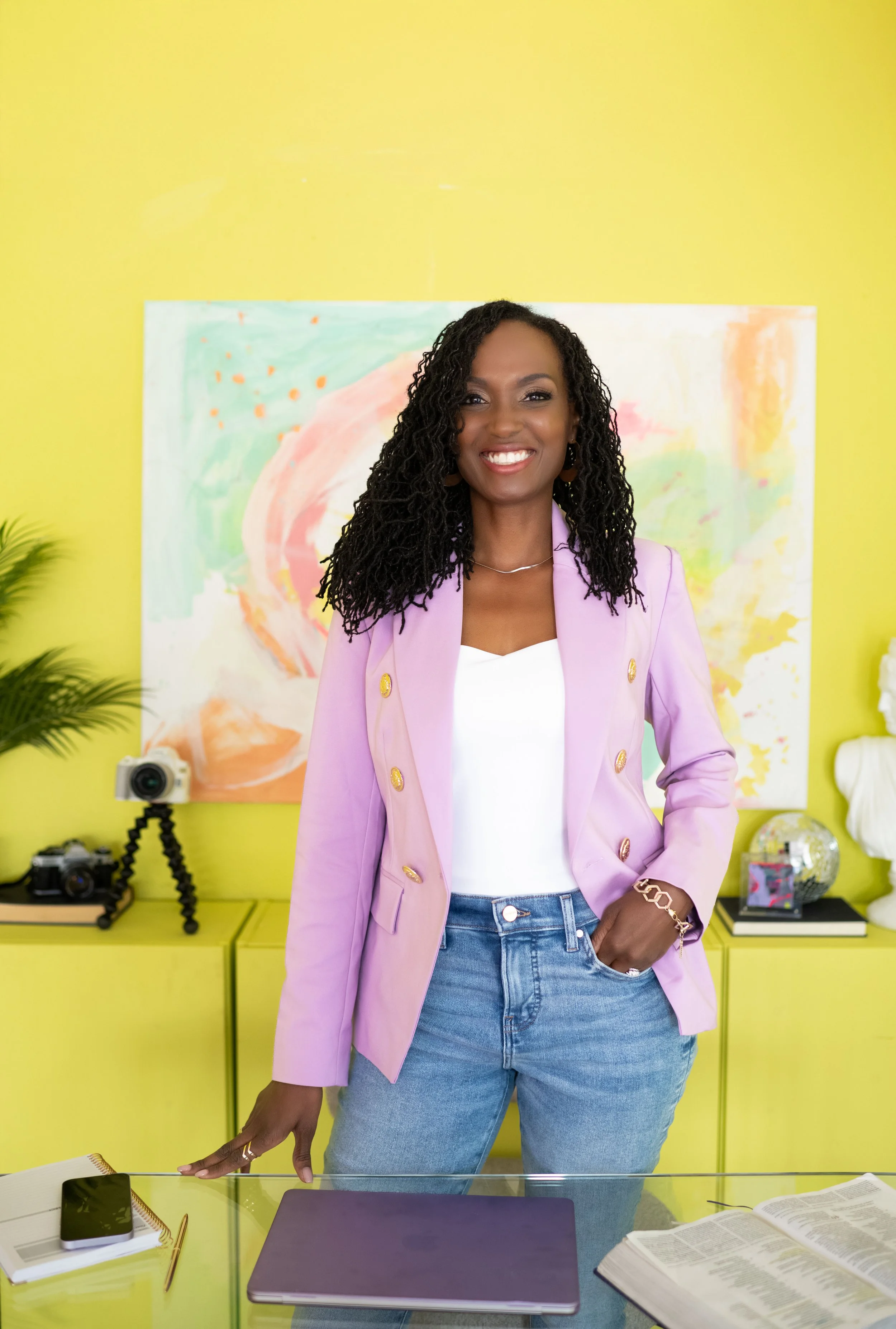 A woman with curly black hair smiling in a colorful room with yellow walls, wearing a pink blazer and jeans, standing behind a glass desk with a laptop, open book, smartphone, and notebook.