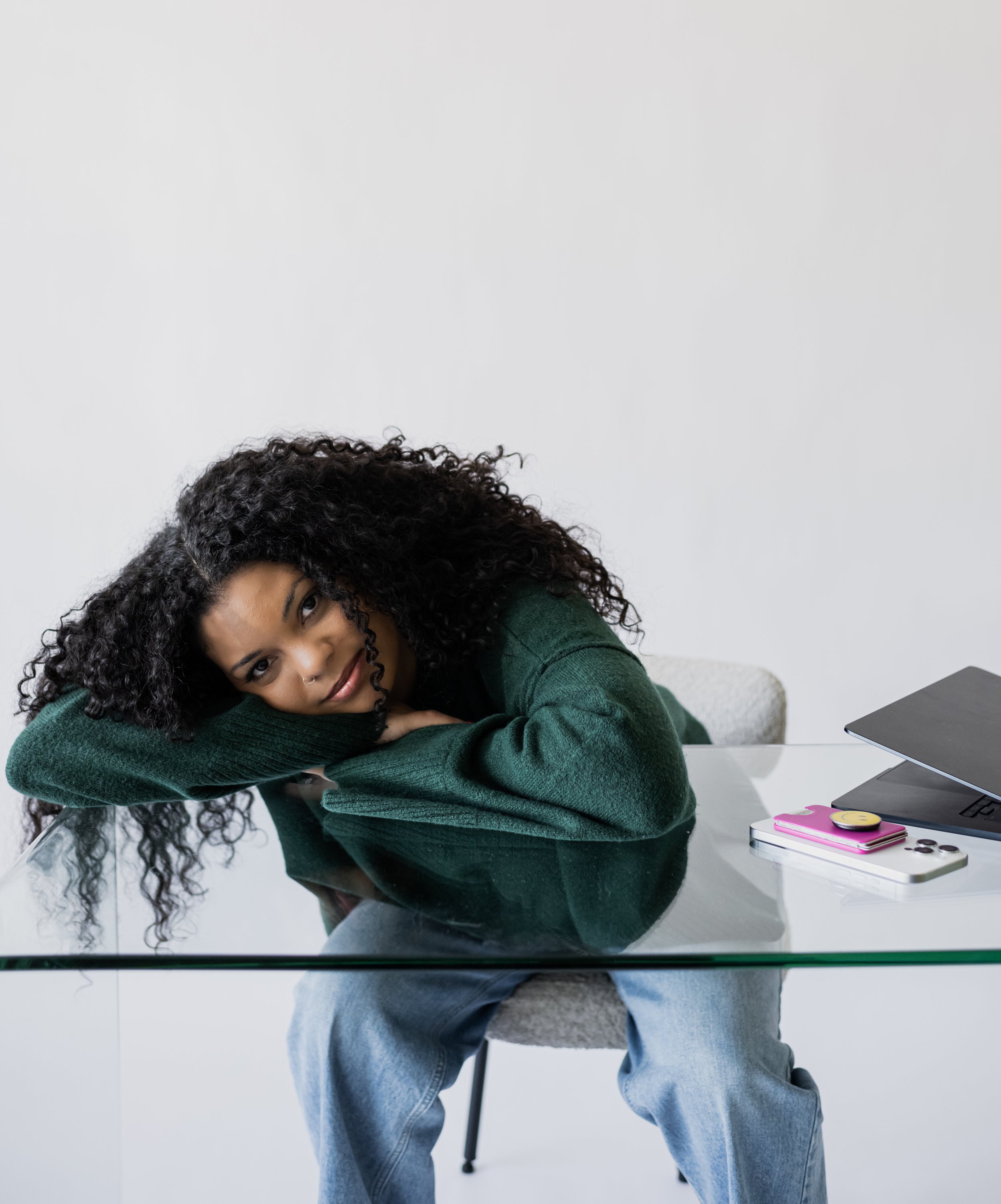Young woman with curly hair sitting at a glass table, resting her head on her arms and looking at the camera. On the table are a laptop, a smartphone, and some accessories.