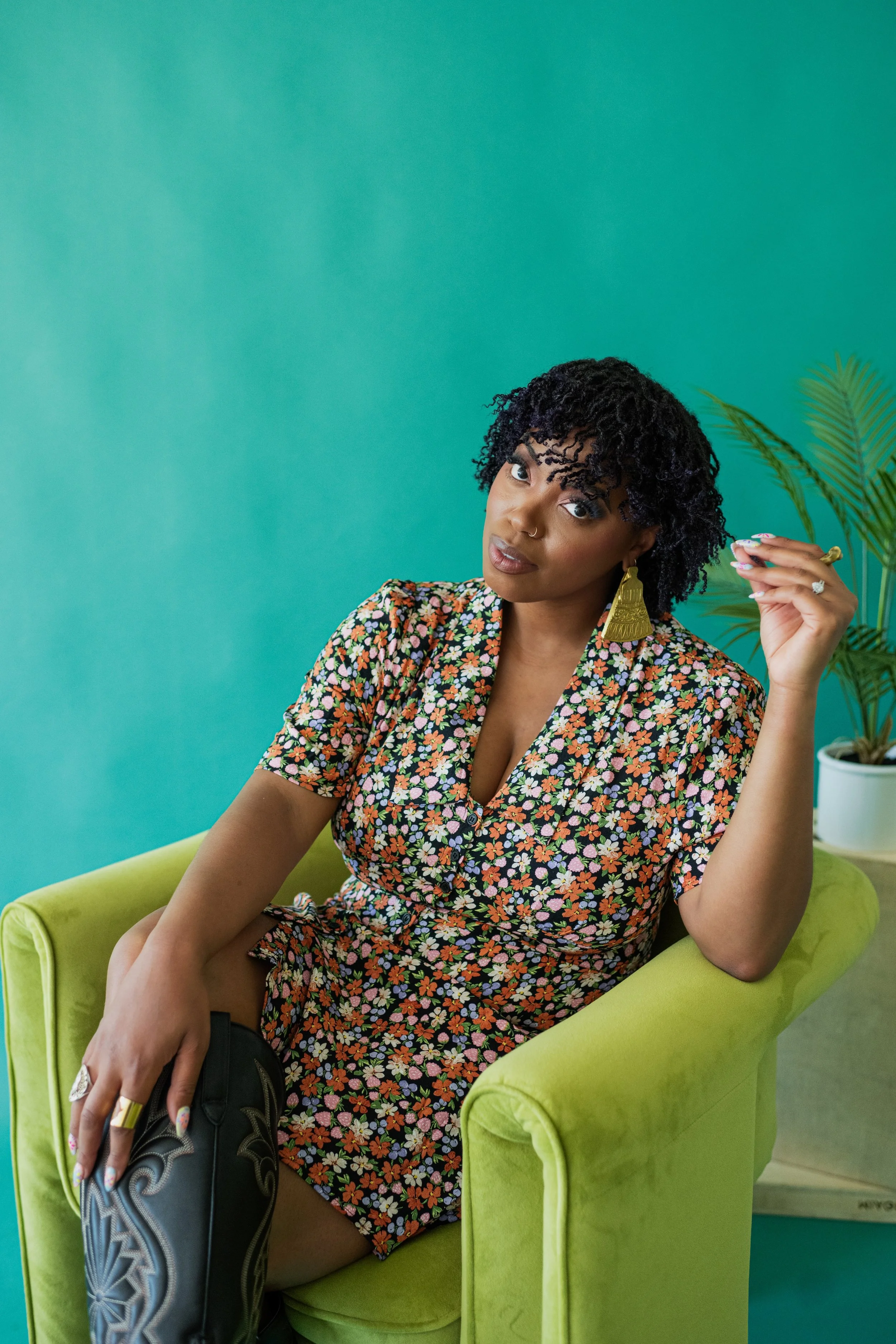 A woman with curly black hair and gold earrings sitting in a lime green armchair against a teal wall with a potted plant in the background.