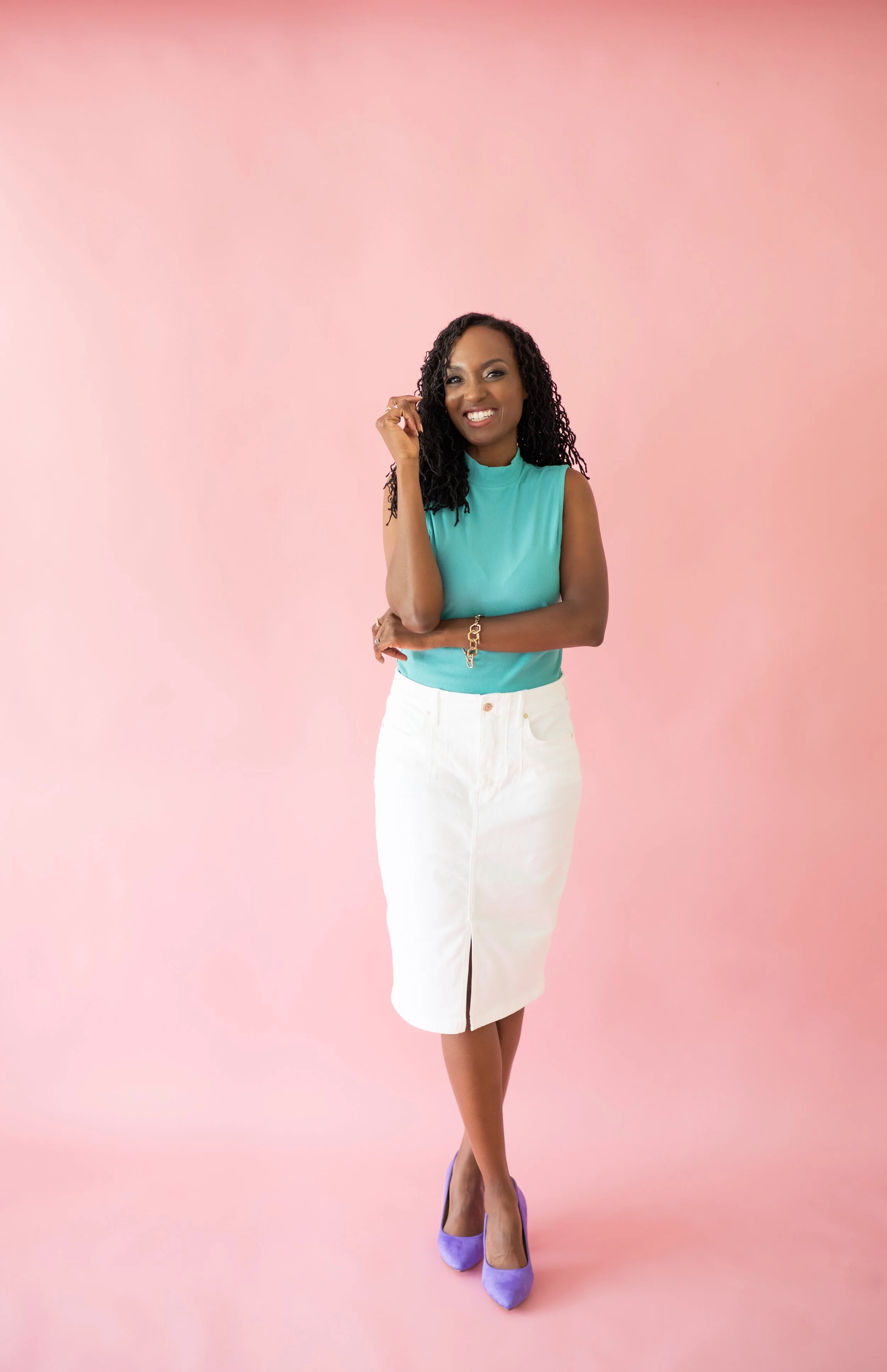A woman with dark curly hair, wearing a sleeveless teal top, white skirt, and purple high heels, standing against a soft pink background, smiling, with her right arm raised and her left arm crossed over her waist.