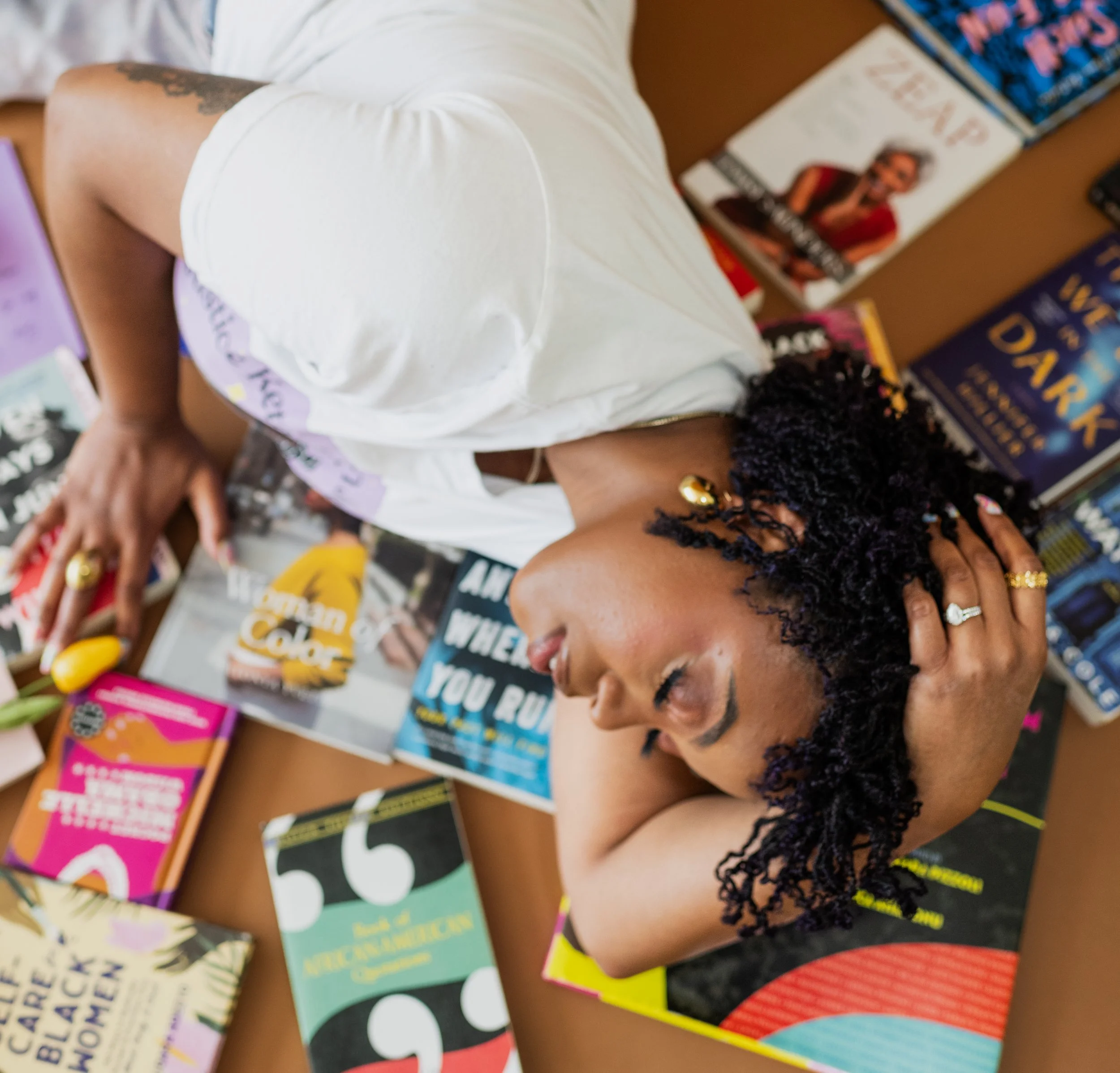 A woman with curly black hair and wearing a white T-shirt is lying on a table covered with colorful books, including titles about humor, relationships, and women's issues, with her hand resting on her head and her eyes closed.
