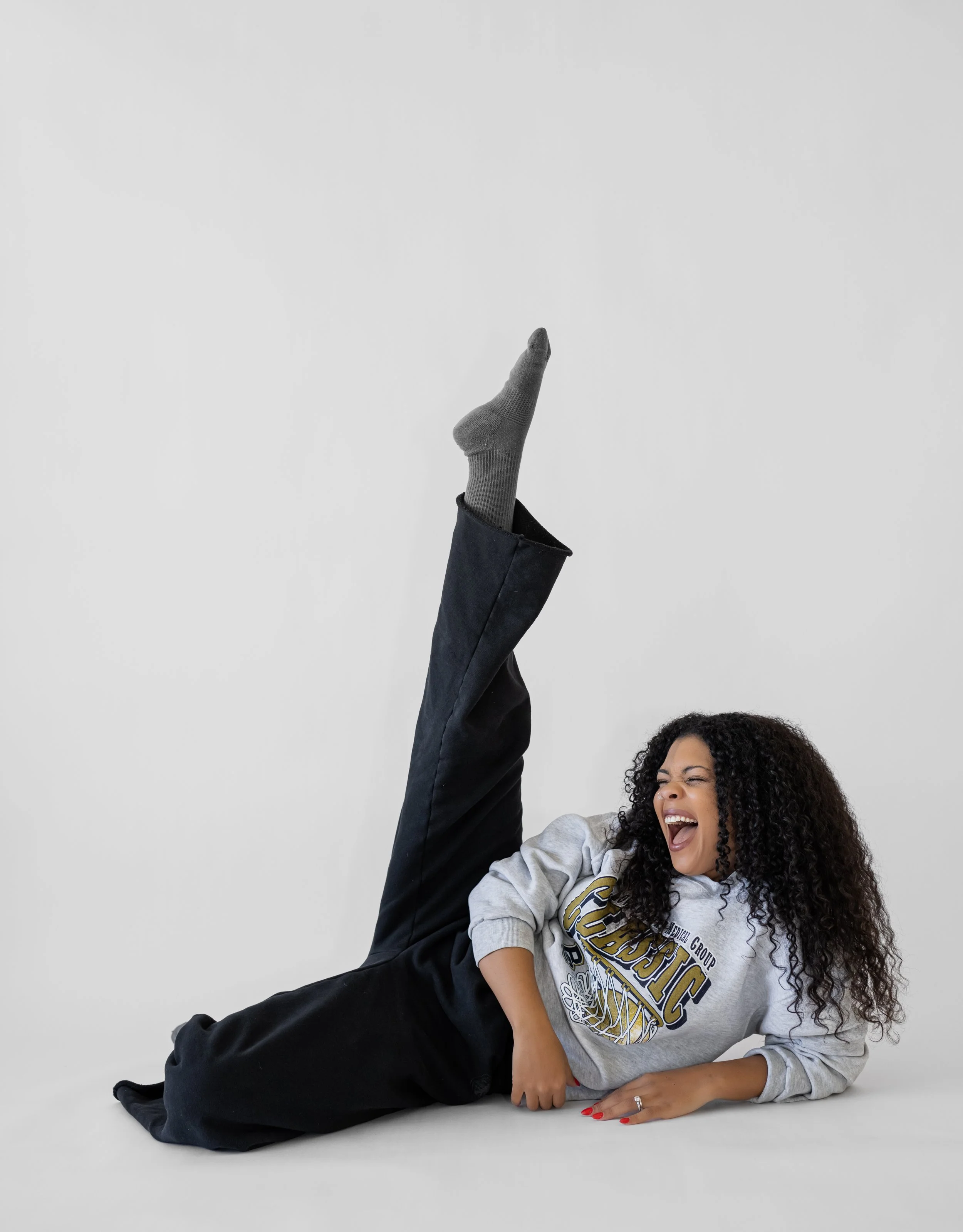 Woman lying on the floor, laughing, with her head turned to the side, wearing a gray sweatshirt and black pants, with her right leg raised in the air, wearing a gray sock.