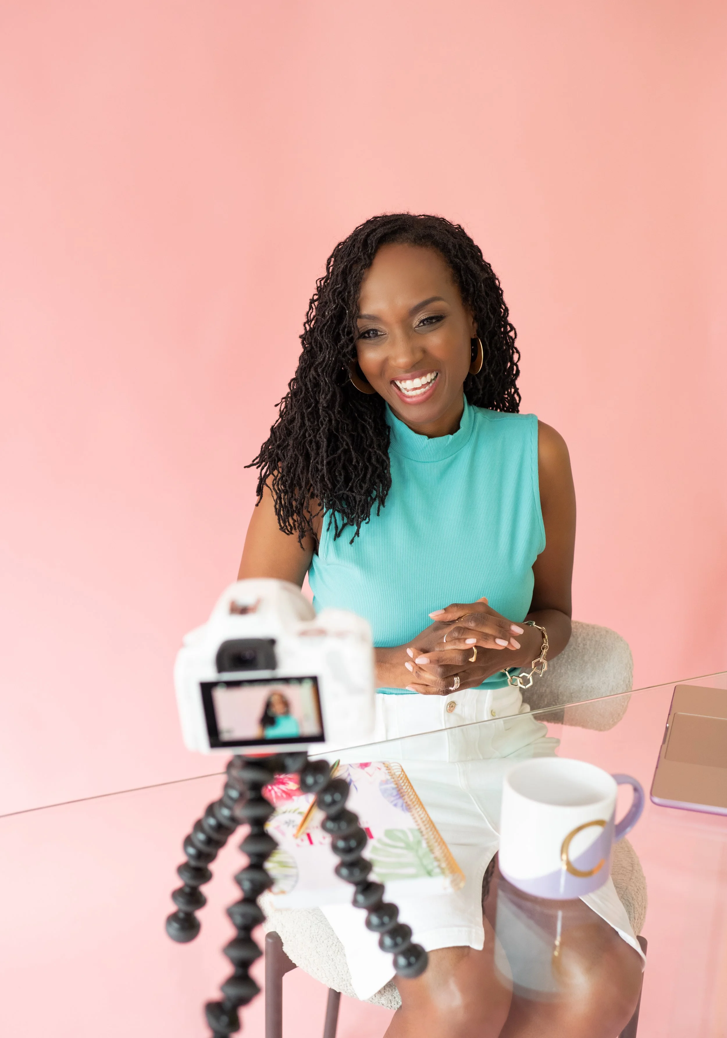A woman with dark curly hair wearing a sleeveless turquoise top and white shorts is sitting at a glass table in front of a pink background. She is smiling and looking at a camera mounted on a flexible tripod, which is positioned on the table. The tab