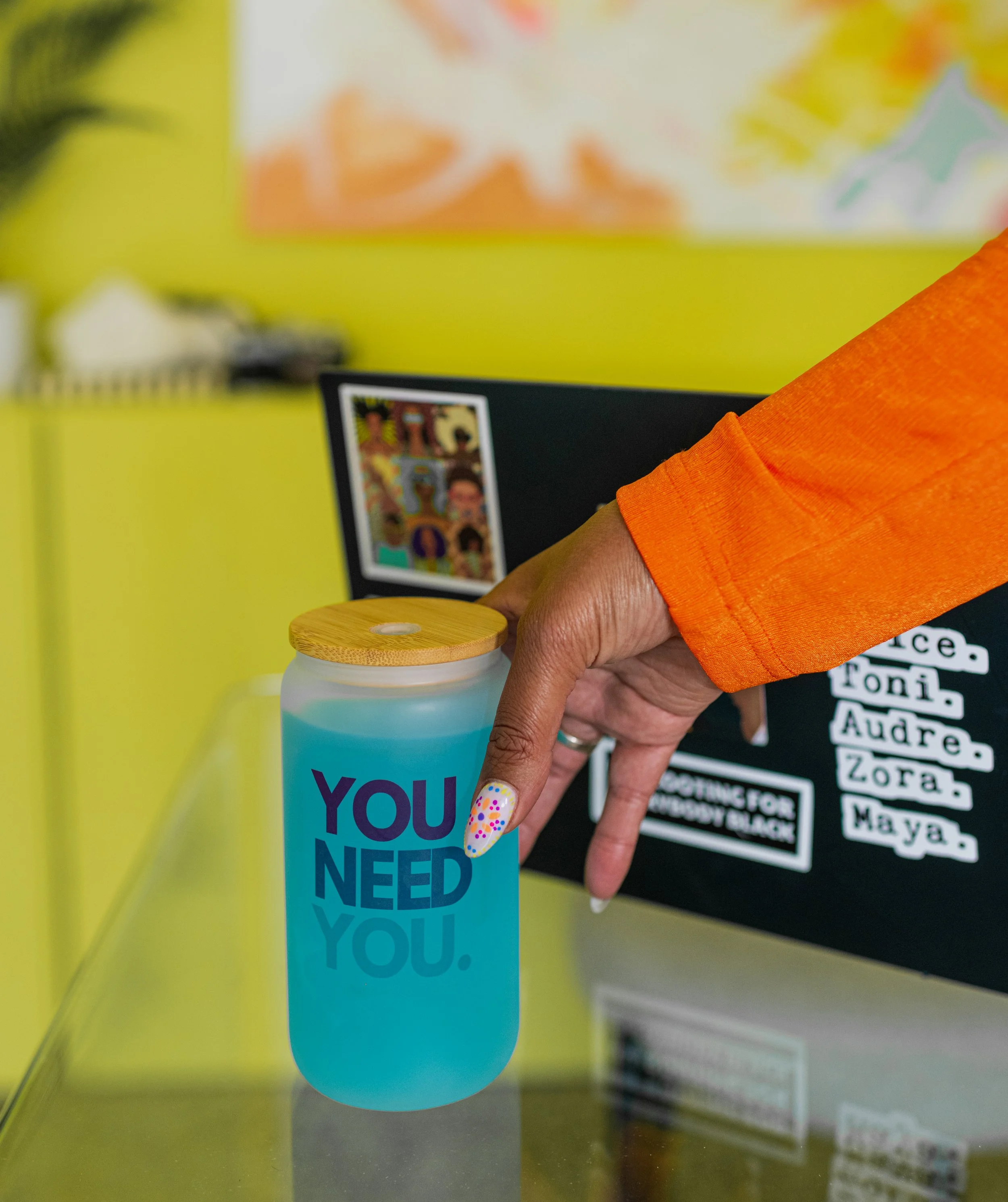 A person in an orange shirt holds a blue tumbler with a wooden lid, saying 'YOU NEED YOU.' in front of a black surface with collage and white text. The background has a lime green wall with blurred artwork.