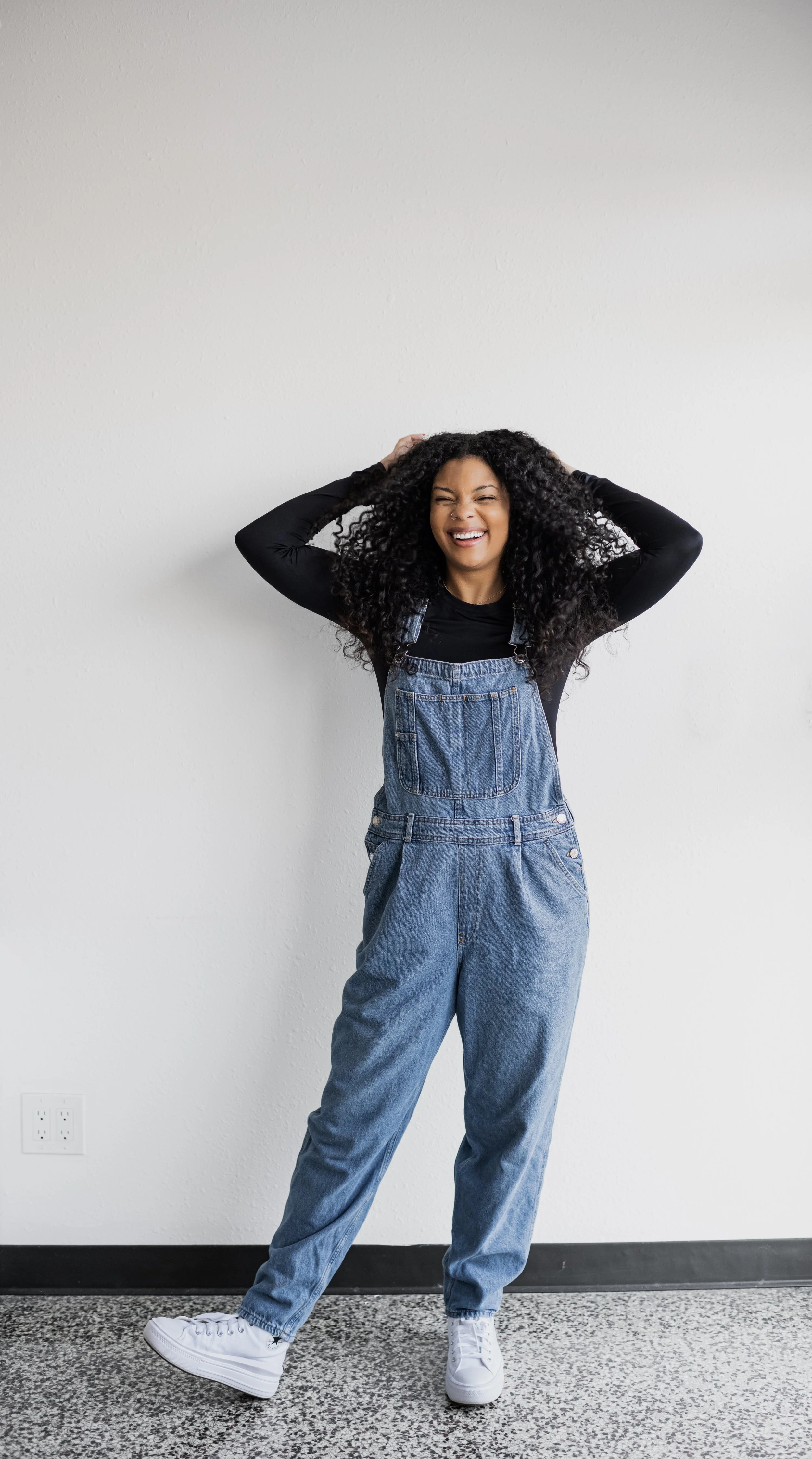 A woman with curly dark hair wearing a black long-sleeve shirt, denim overalls, and white sneakers, standing against a plain white wall, smiling with eyes closed and hands on her head.