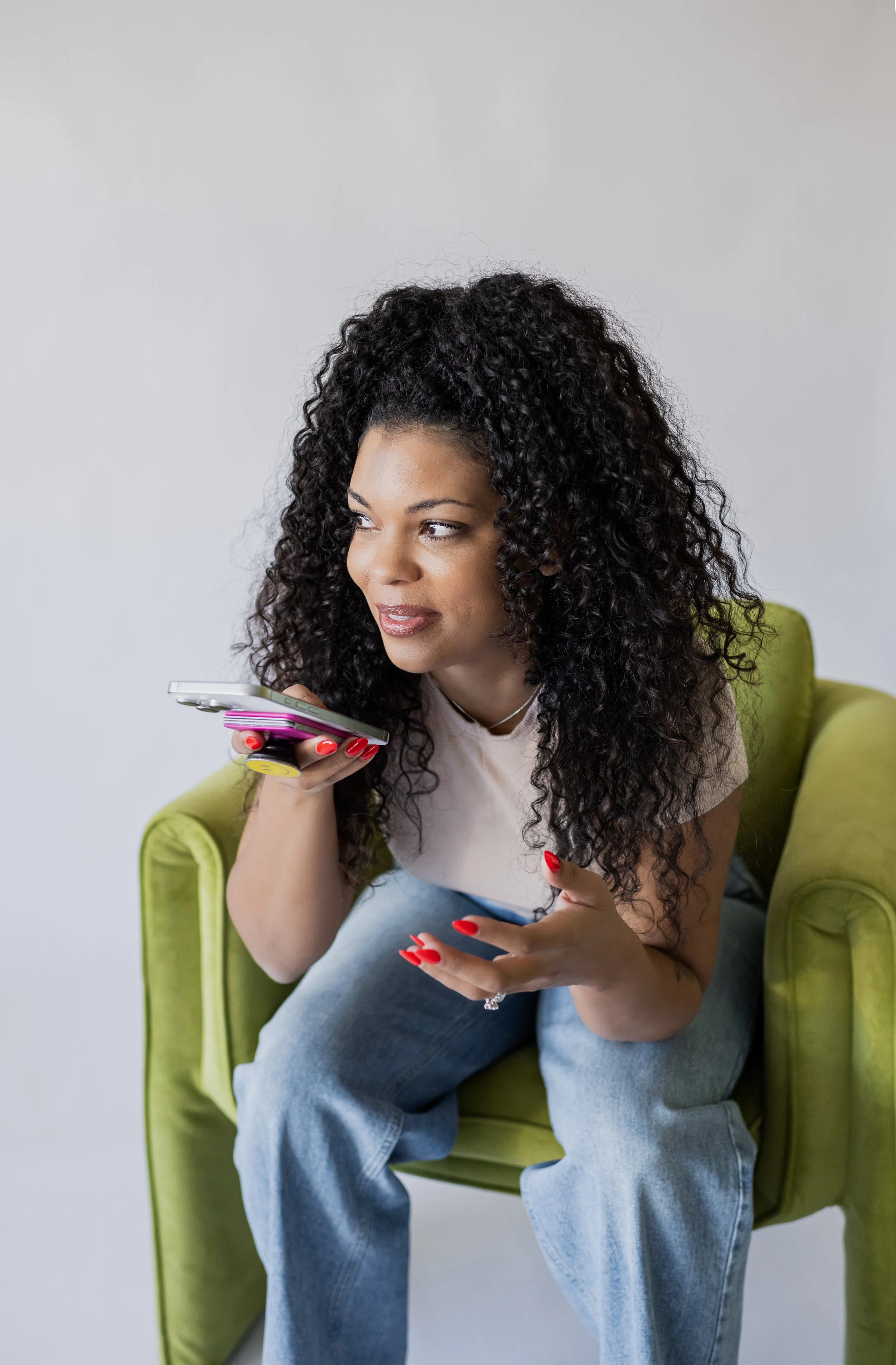 A woman with curly black hair, wearing a white t-shirt and blue jeans, sitting on a green armchair, holding a smartphone with a pink pop socket to her ear, looking to the side with a slight smile.
