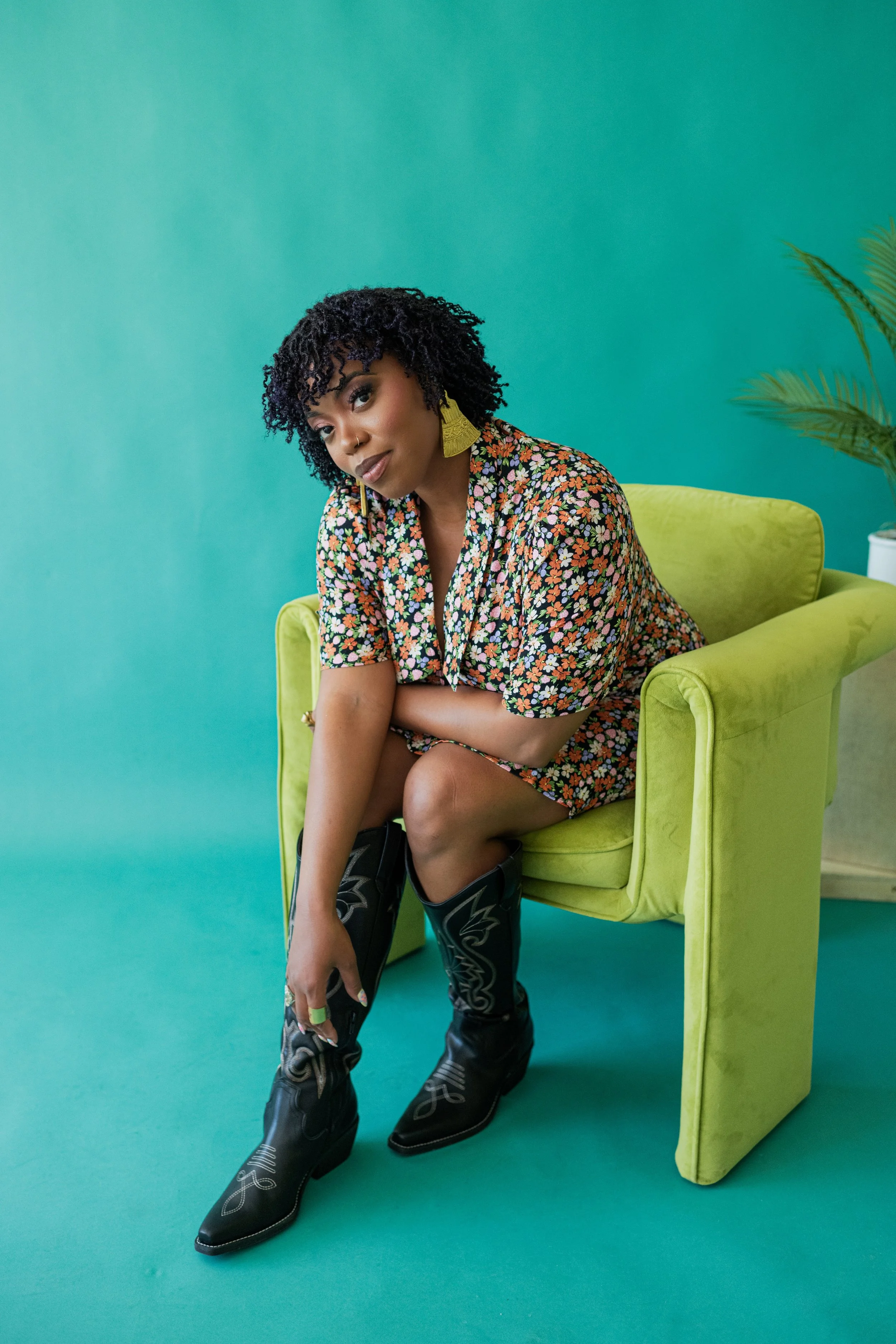 A woman with curly black hair sitting on a green armchair against a teal background, wearing a colorful floral romper, black cowboy boots, and large yellow earrings.