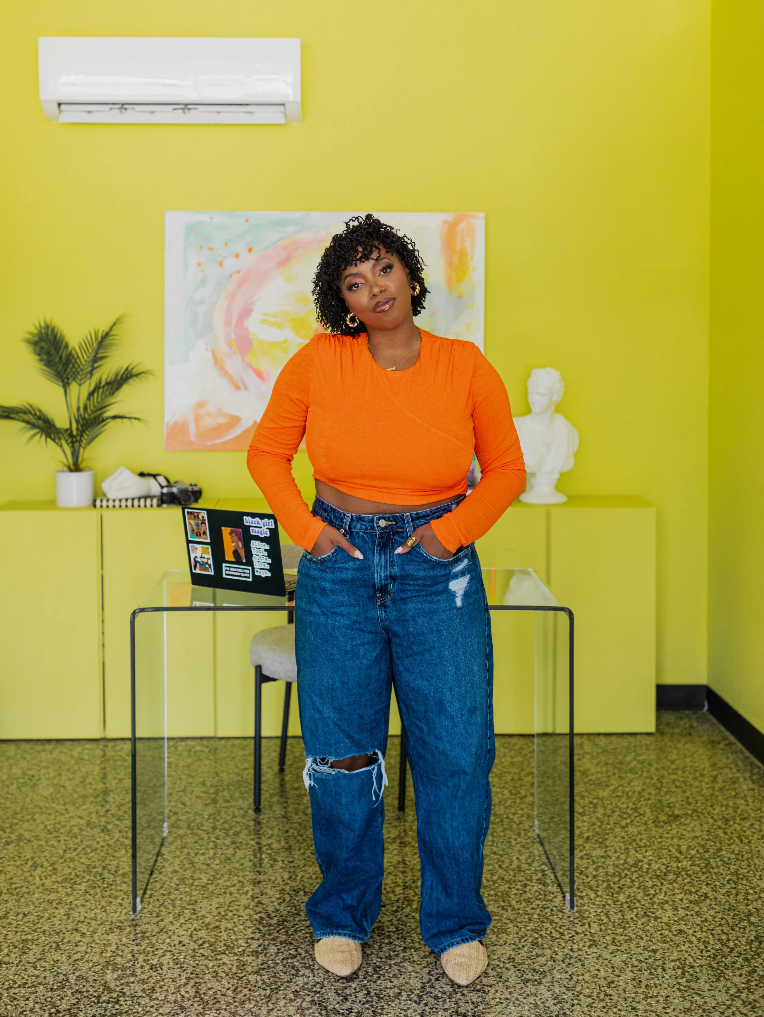 A woman with curly hair wearing an orange crop top and ripped blue jeans stands with hands in pockets in a brightly colored room with lime green walls, abstract art, a white bust, a potted plant, and a desk with a laptop.