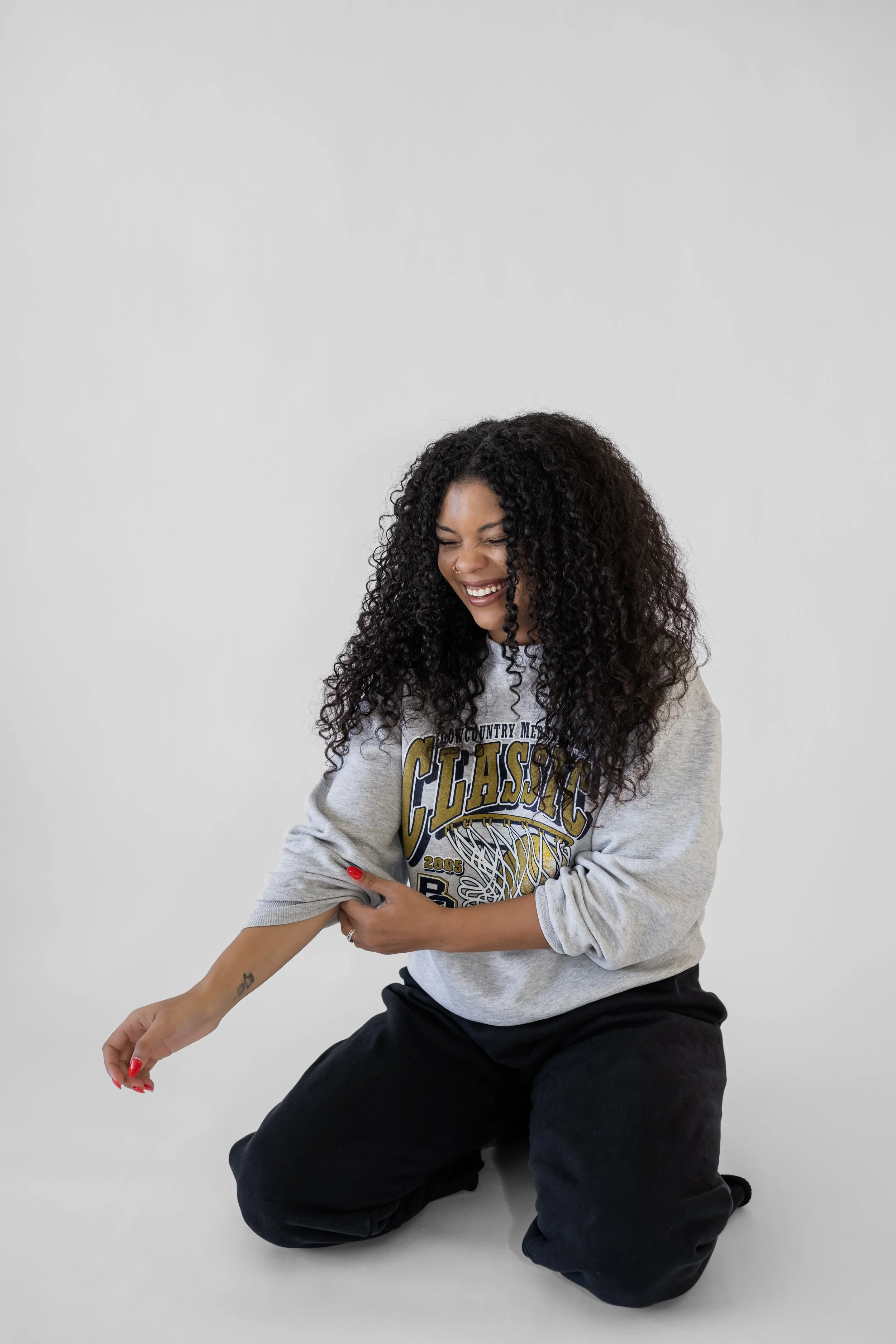Woman with curly hair smiling, kneeling on the floor, wearing a gray sweatshirt and black pants, against a plain white background.